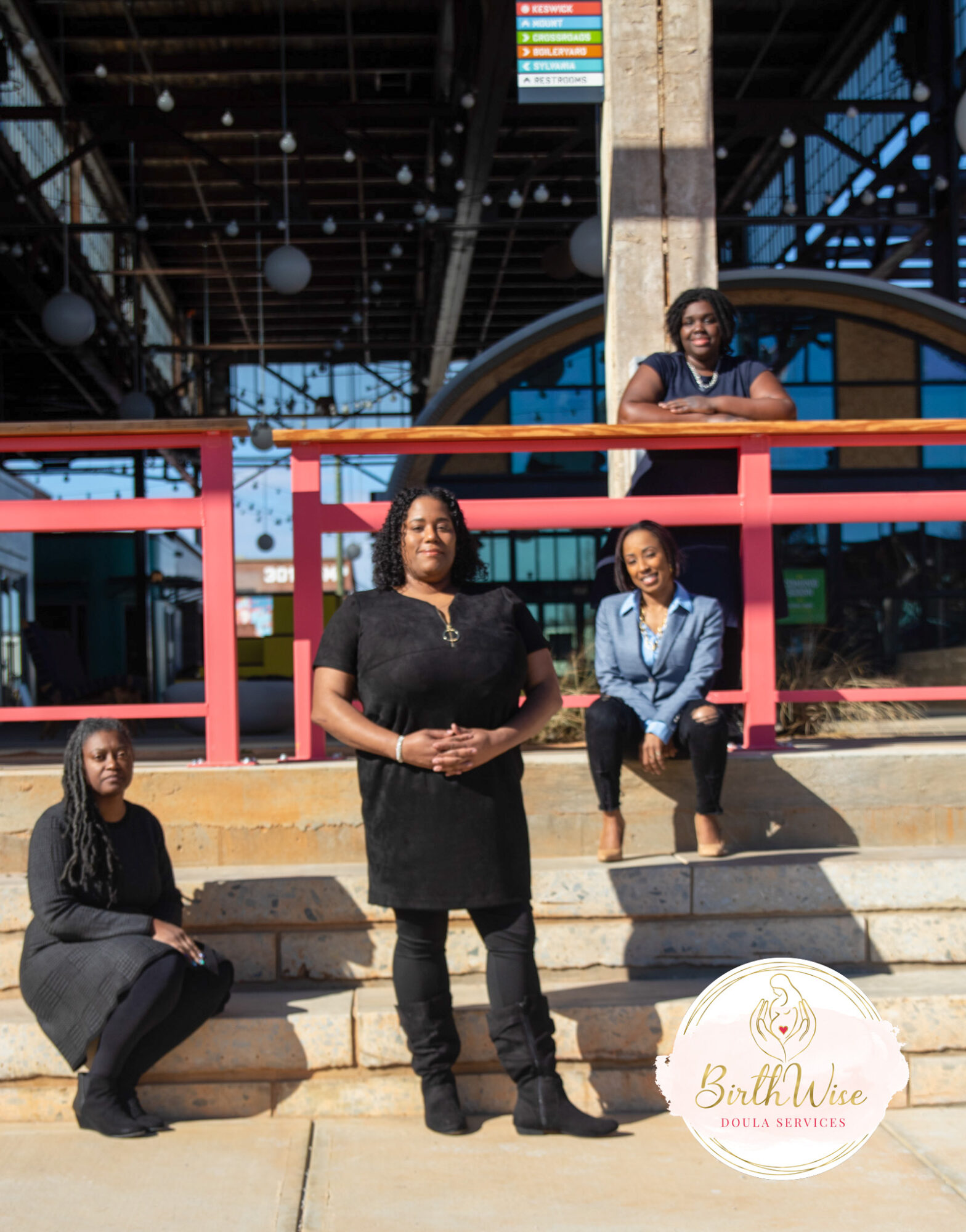 Four women pose outdoors in front of a modern building with glass and metal architecture, some sitting and some standing.