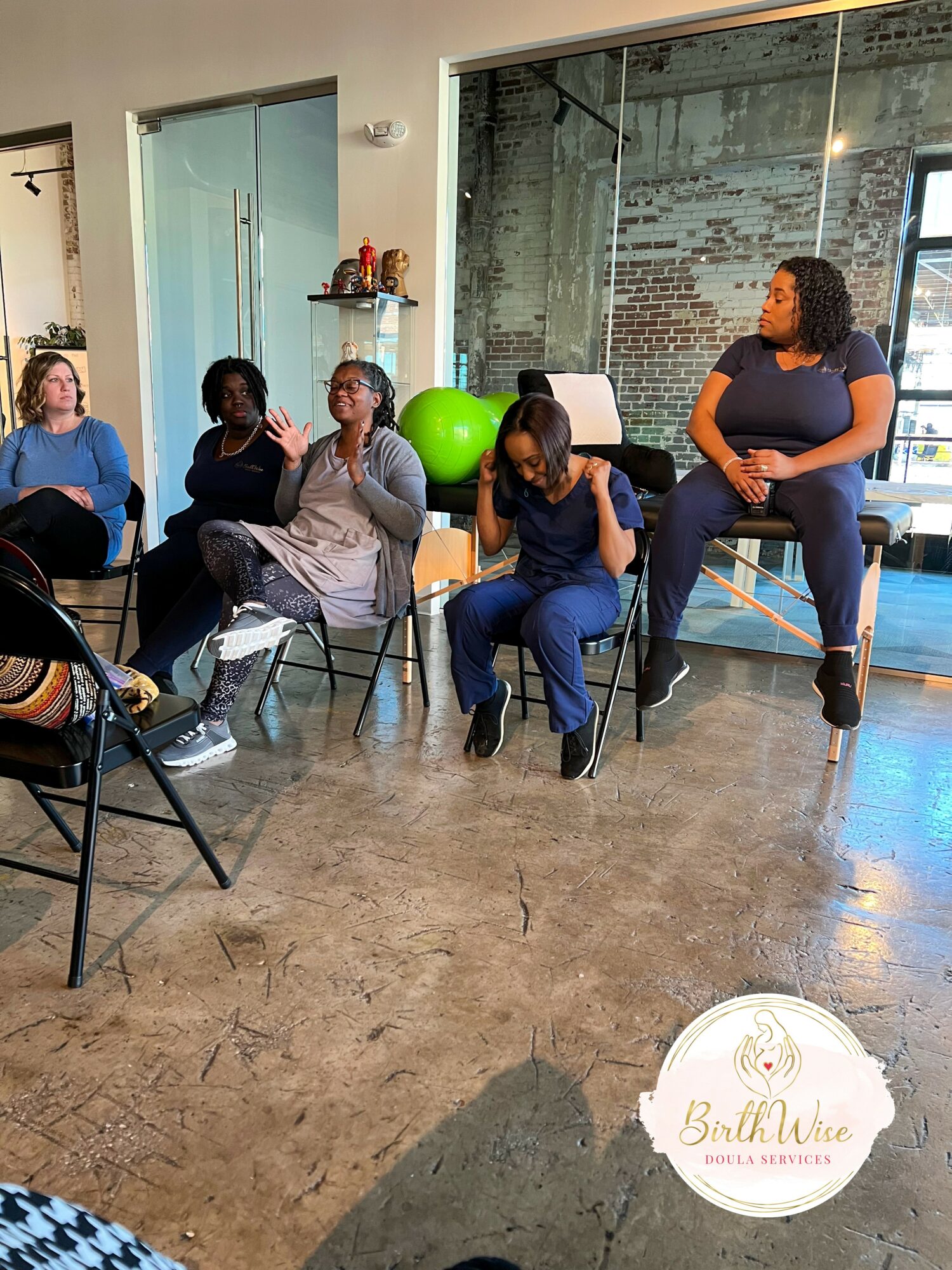 Group of five women seated and standing in a room with large windows and brick wall, engaged in conversation.