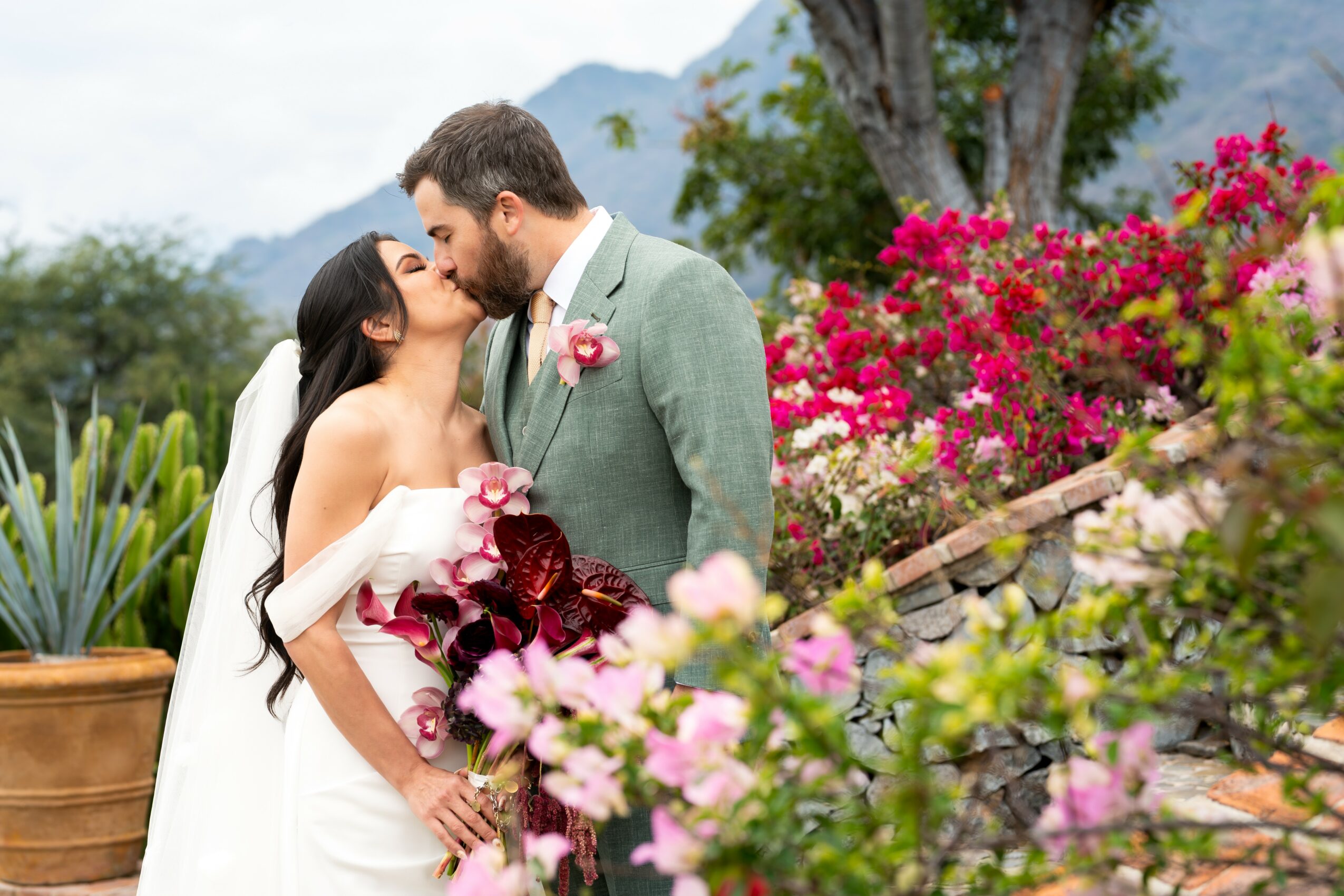 Bride and groom share a kiss outdoors with colorful flowers and mountains in the background.