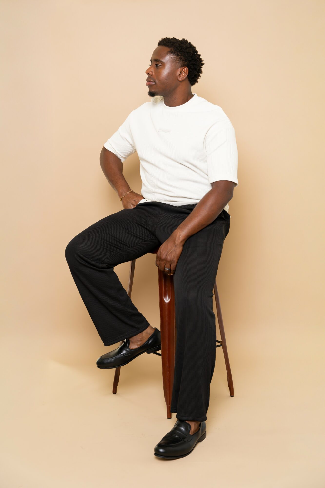Young man sitting on a wooden stool against a beige background, wearing a white t-shirt and black pants.