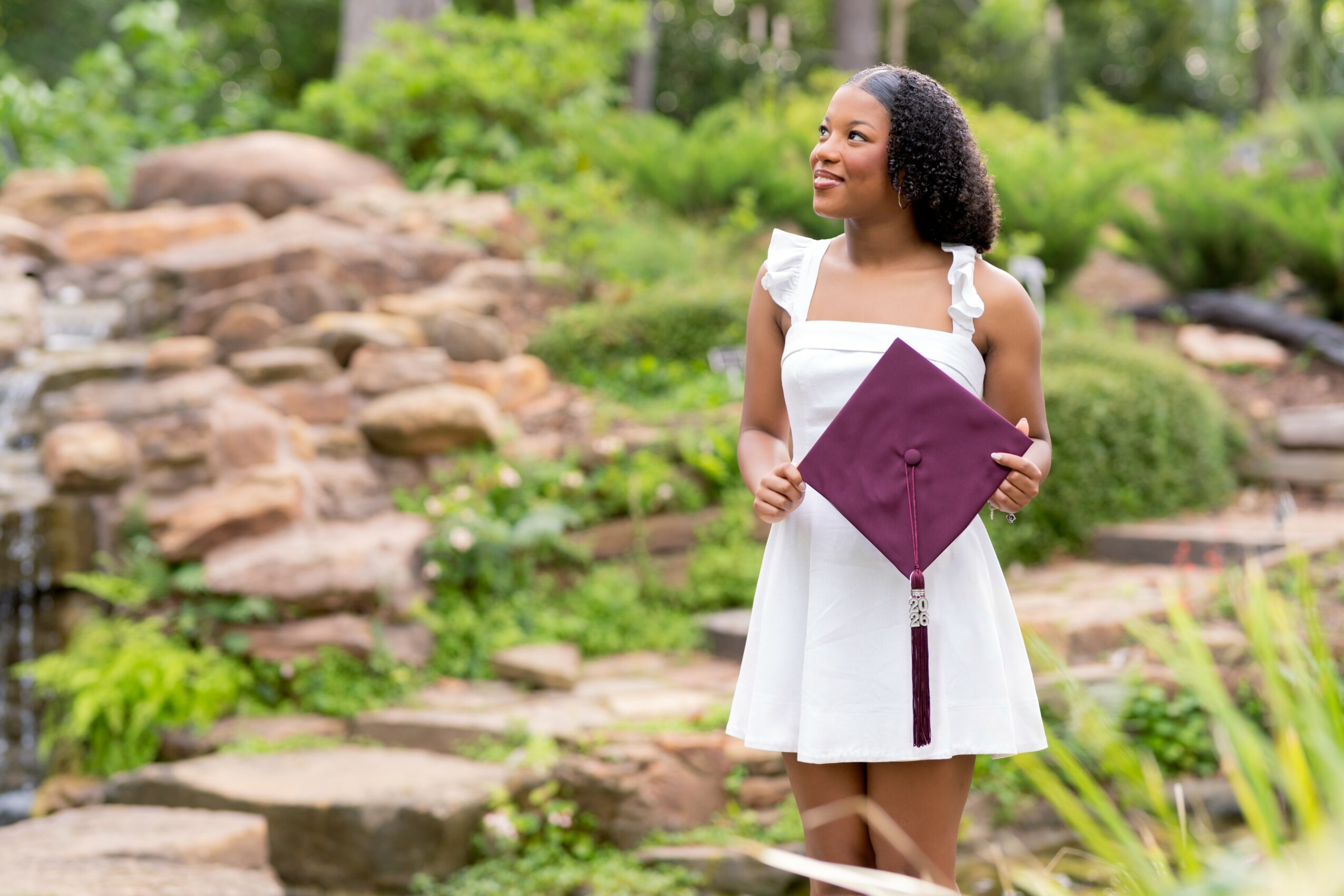 Young woman in white dress holding a maroon folder outdoors with rocks and greenery.