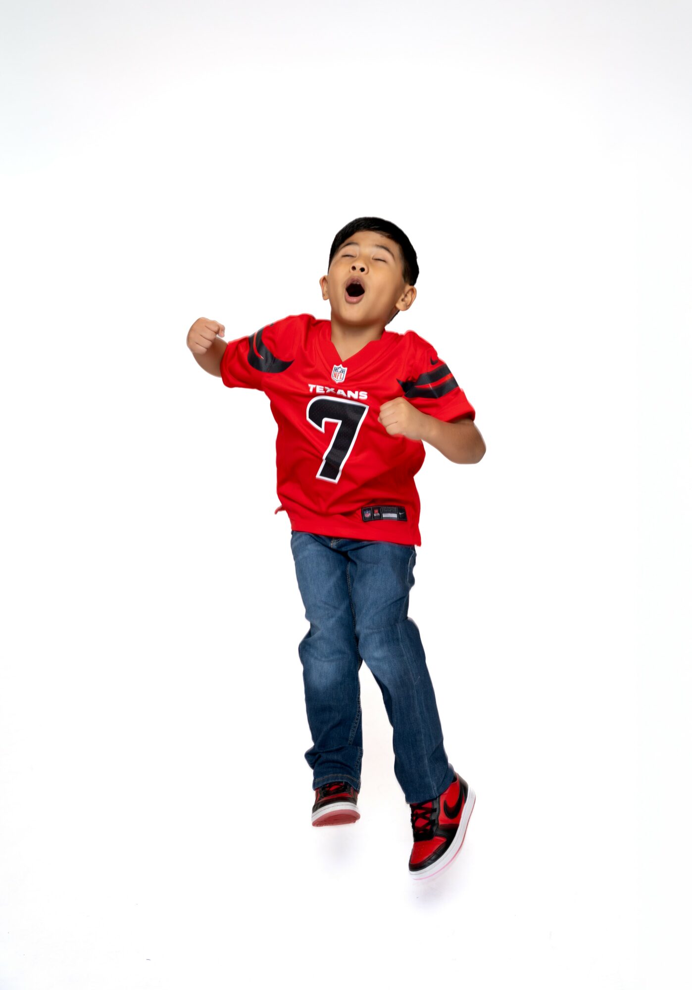 Child wearing a red sports jersey and jeans, jumping with arms raised and mouth open.