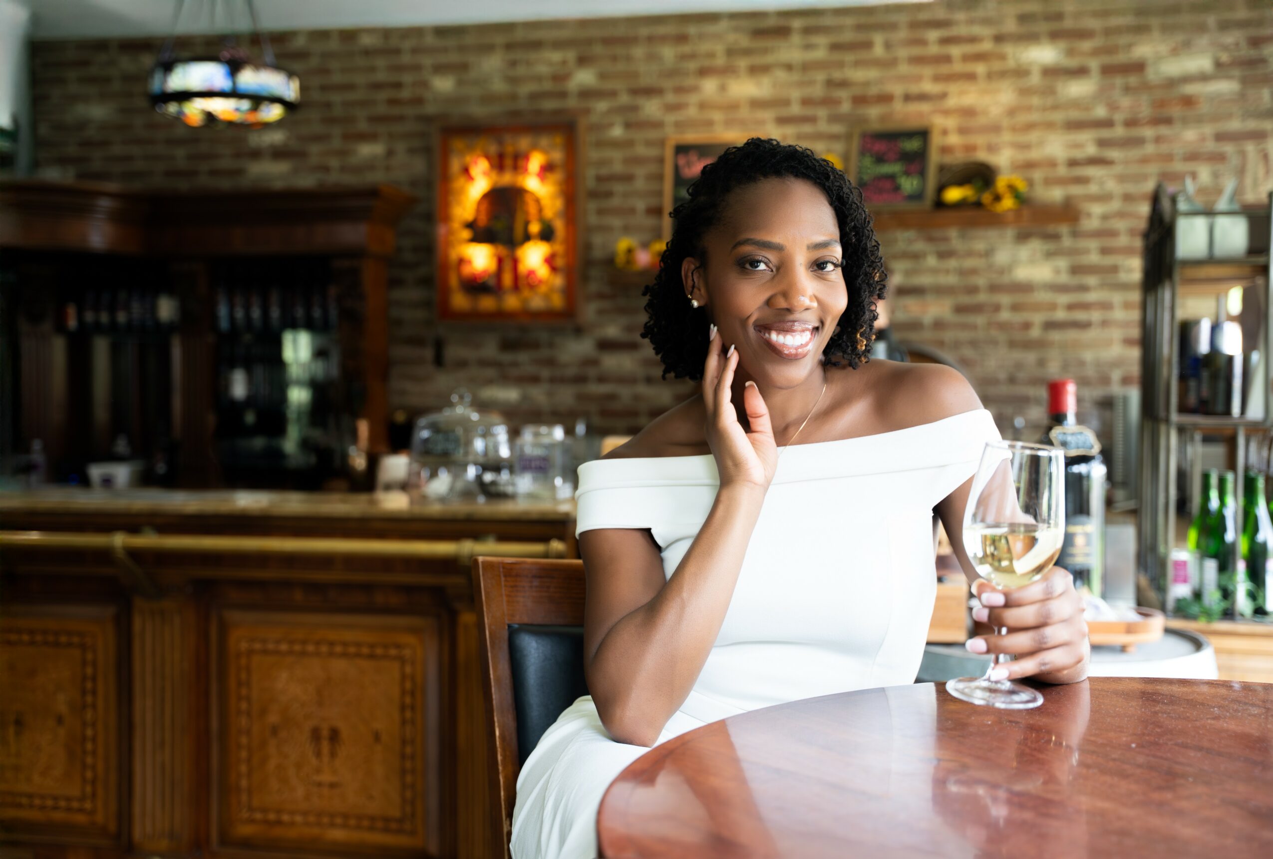Woman in white dress sitting at bar with glass of white wine, smiling, in a cozy restaurant setting.