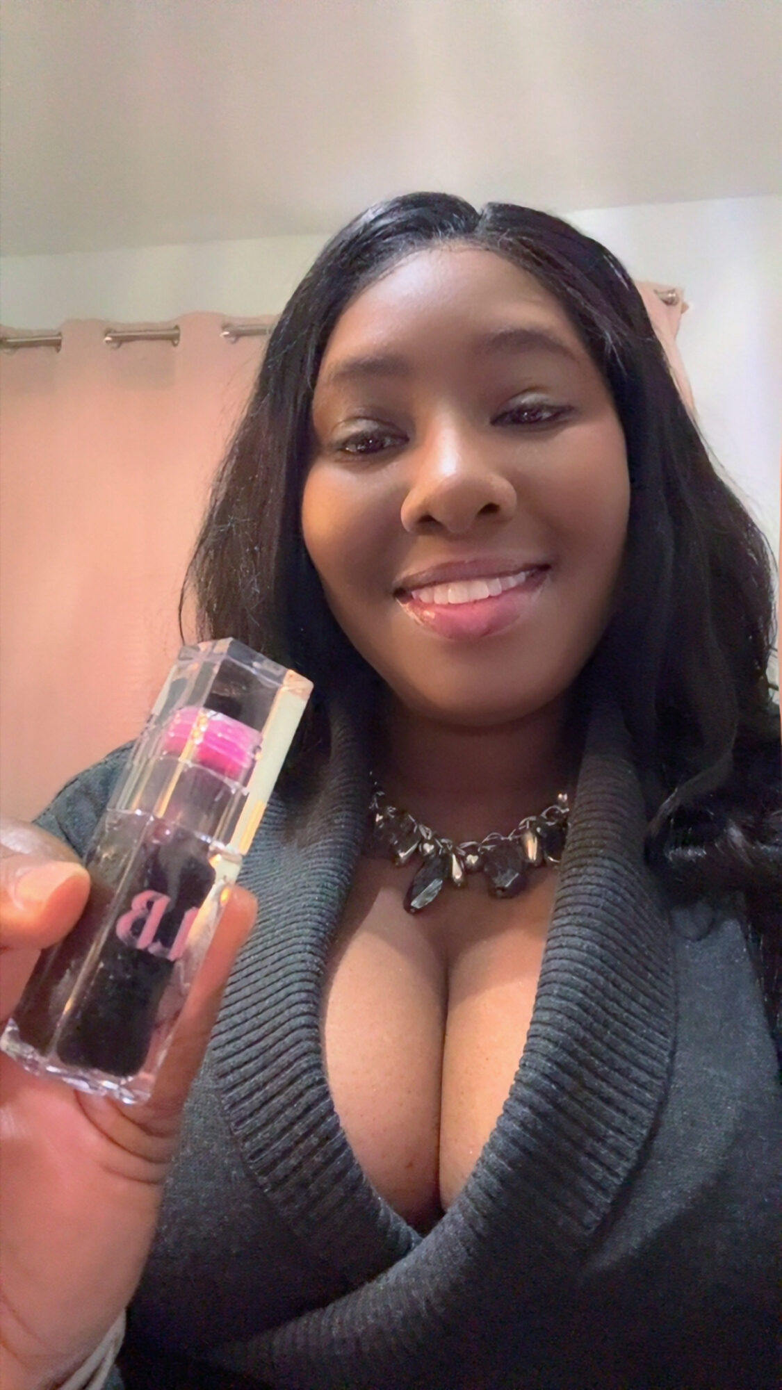 Woman with dark hair smiling, holding a lipstick, wearing a necklace and a dark top with a deep neckline.