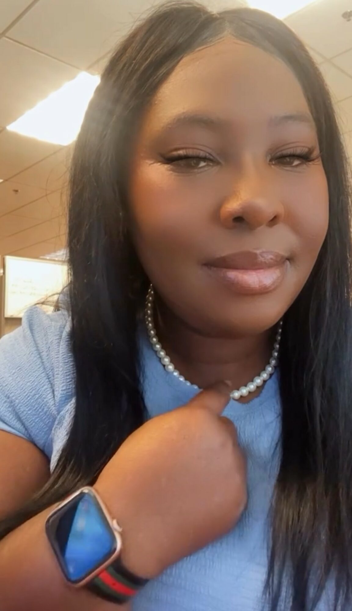 Woman with long dark hair wearing a blue top, pearl necklace, and smartwatch, indoors with warm lighting.