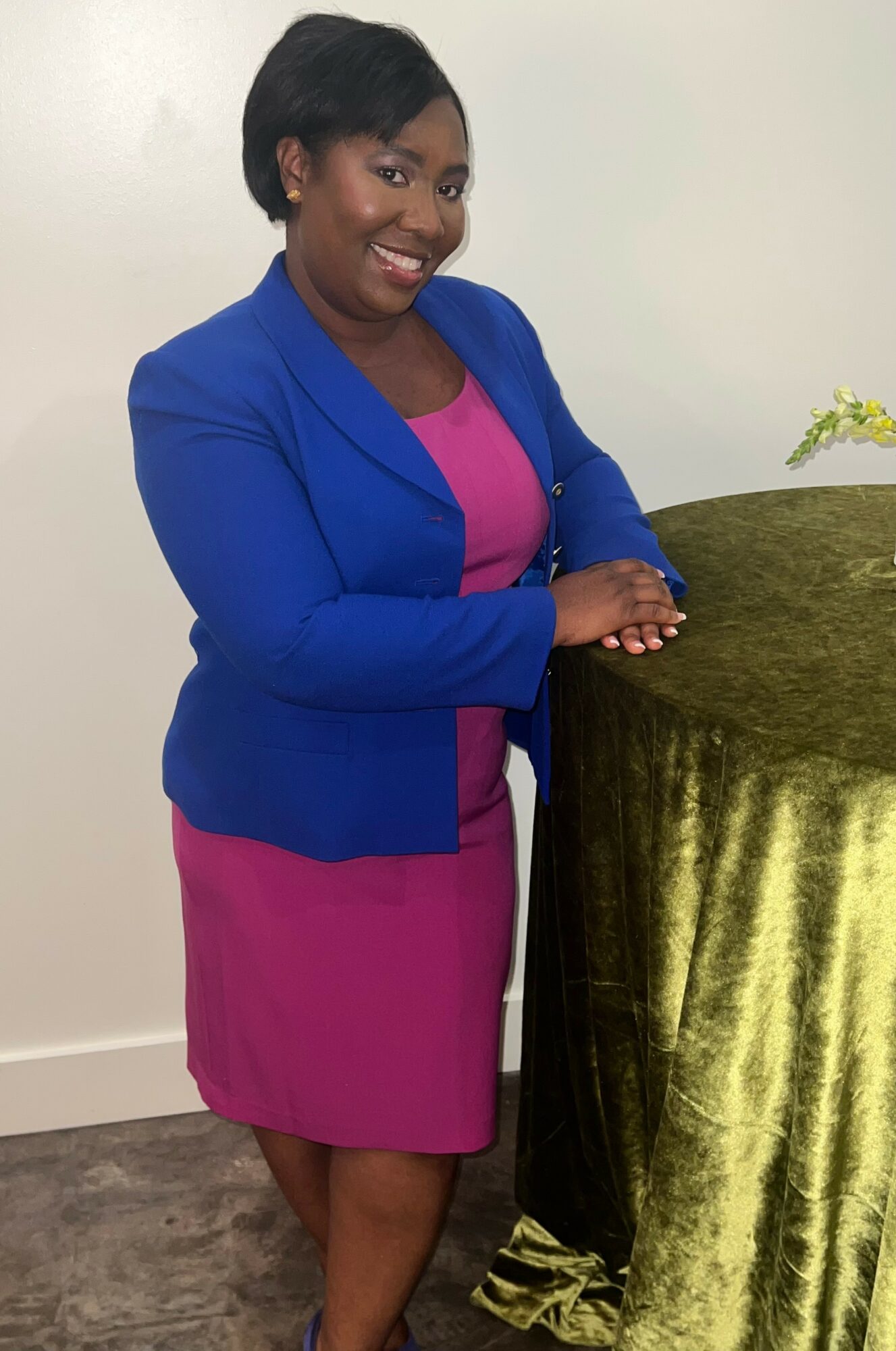 Woman in blue blazer and pink dress standing beside a table with green cloth, smiling at camera.