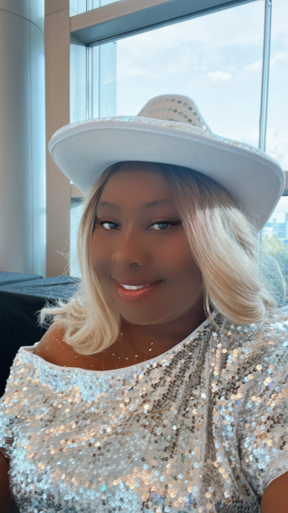 Woman with blonde hair wearing a large white hat and a sequined top, smiling indoors near a window.