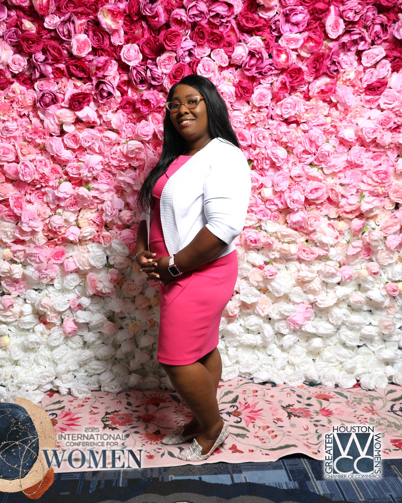 Woman in pink dress and white blazer standing in front of a pink and white floral wall, smiling.