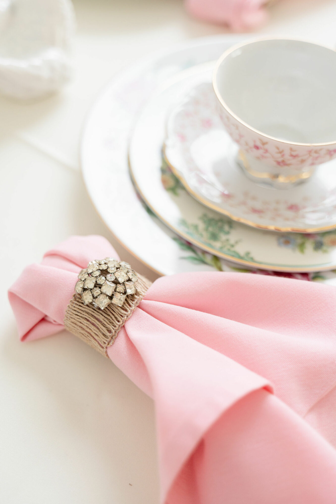 Stacked floral-patterned plates with a teacup on top, pink napkin with a decorative napkin ring, on a white surface.