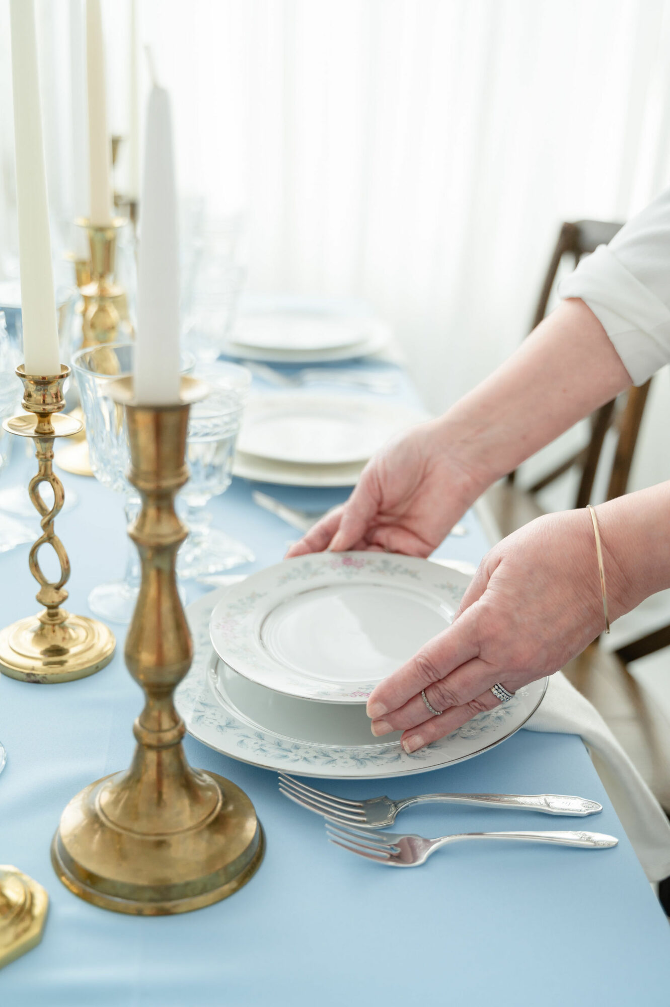 Person stacking white plates on a dining table with blue tablecloth, surrounded by gold candlesticks and tableware.