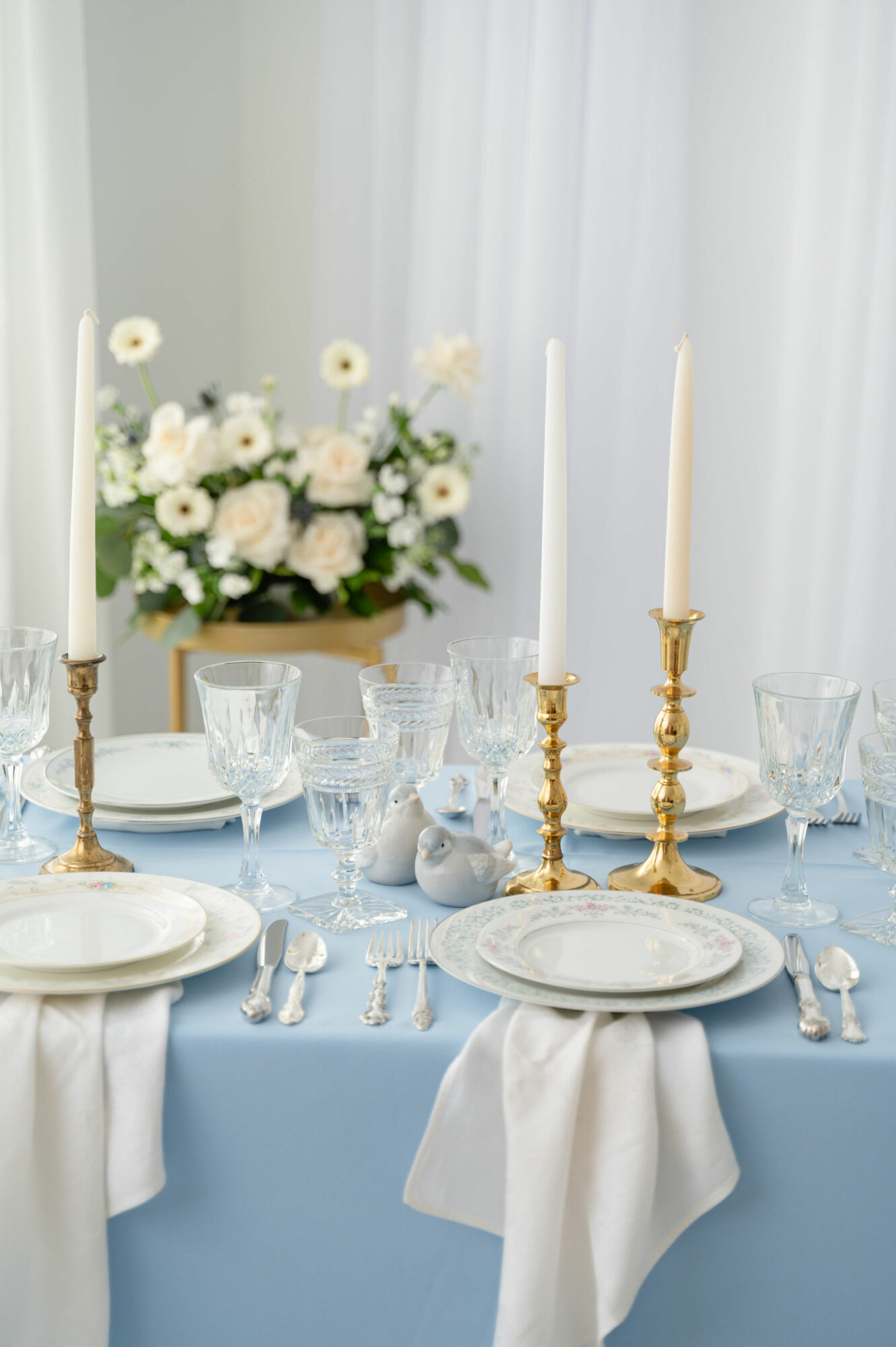 Elegant table setting with white plates, silverware, glassware, and tall candles in gold holders, with a floral centerpiece in the background.
