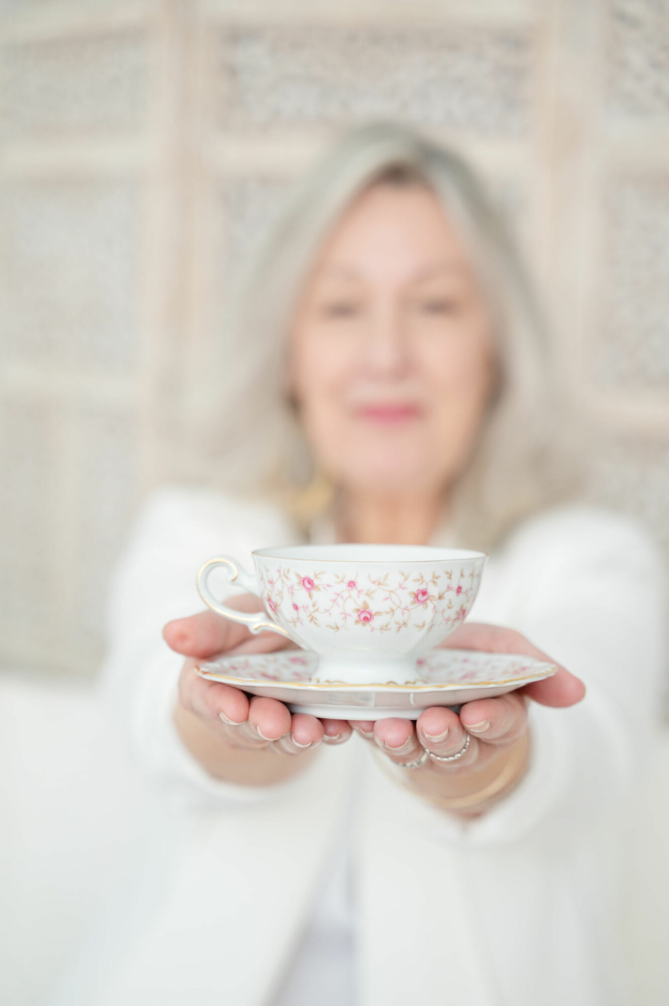 Person holding a floral teacup and saucer, smiling, with a blurred background.