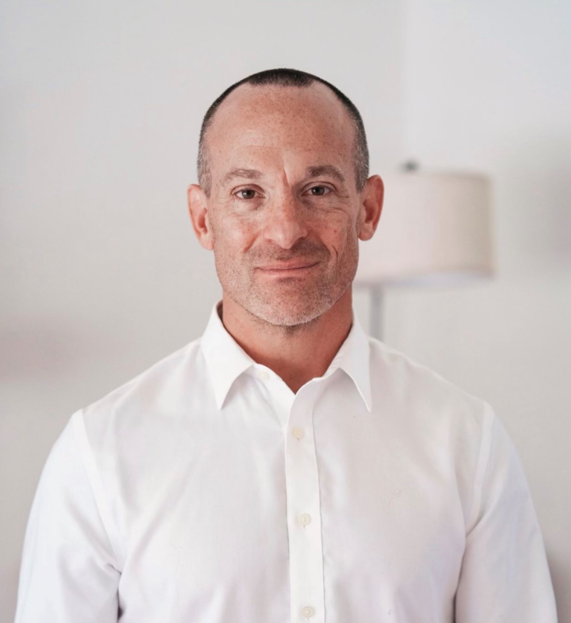 Man with short hair and a slight smile, wearing a white shirt, standing indoors with a lamp in the background.