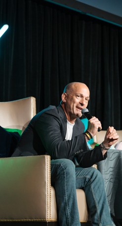 Bald man speaking into a microphone on stage, seated in a beige chair with black curtains behind him.
