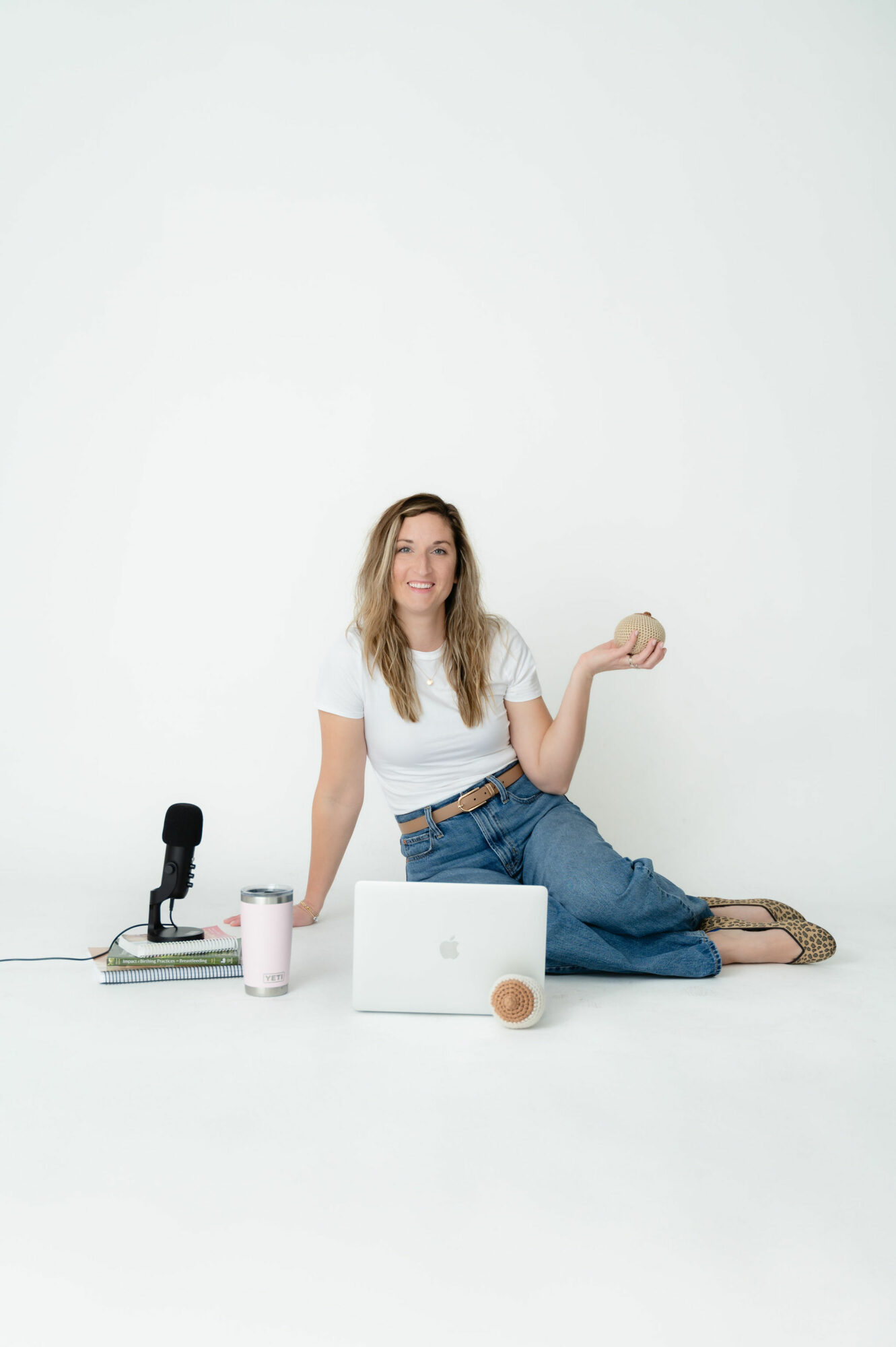 Woman sitting on the floor with a laptop, microphone, and cup, holding a phone in her right hand, against a plain white background.