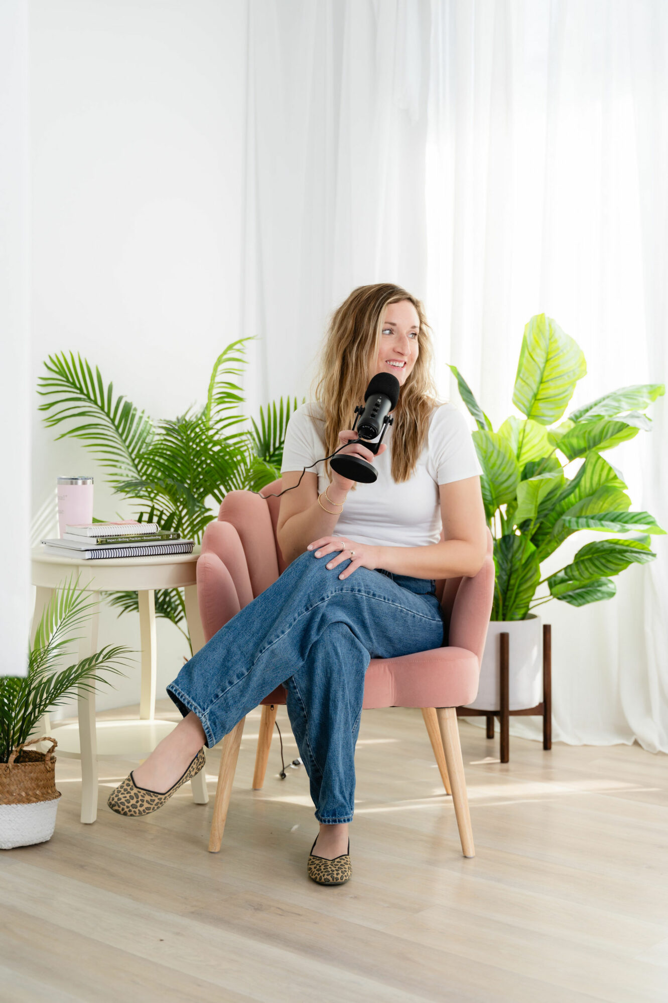 Woman sitting in pink chair holding a camera, surrounded by green plants and white curtains.