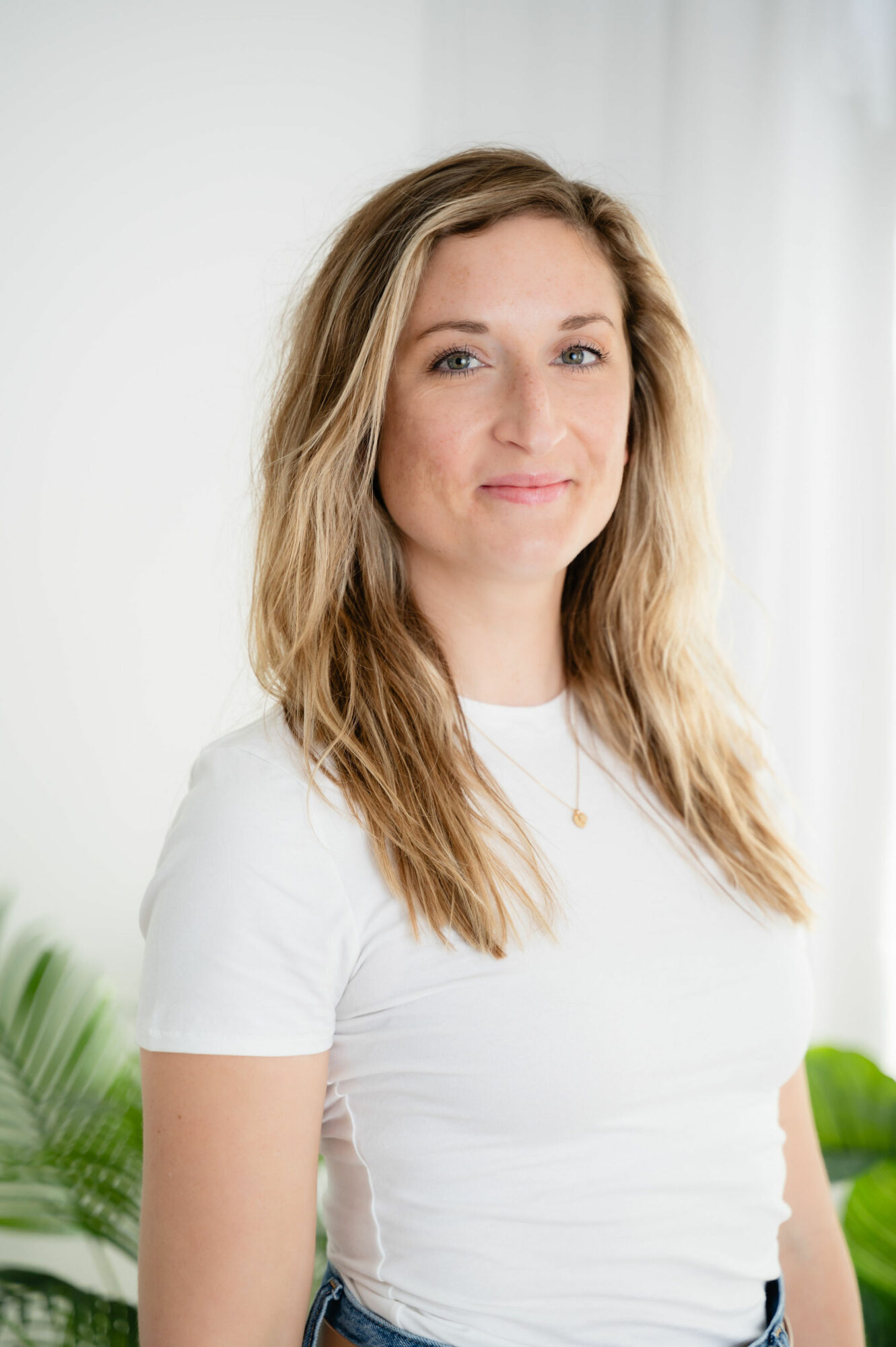 Woman with long wavy hair wearing a white t-shirt, smiling softly, standing indoors with white curtains and green plants.