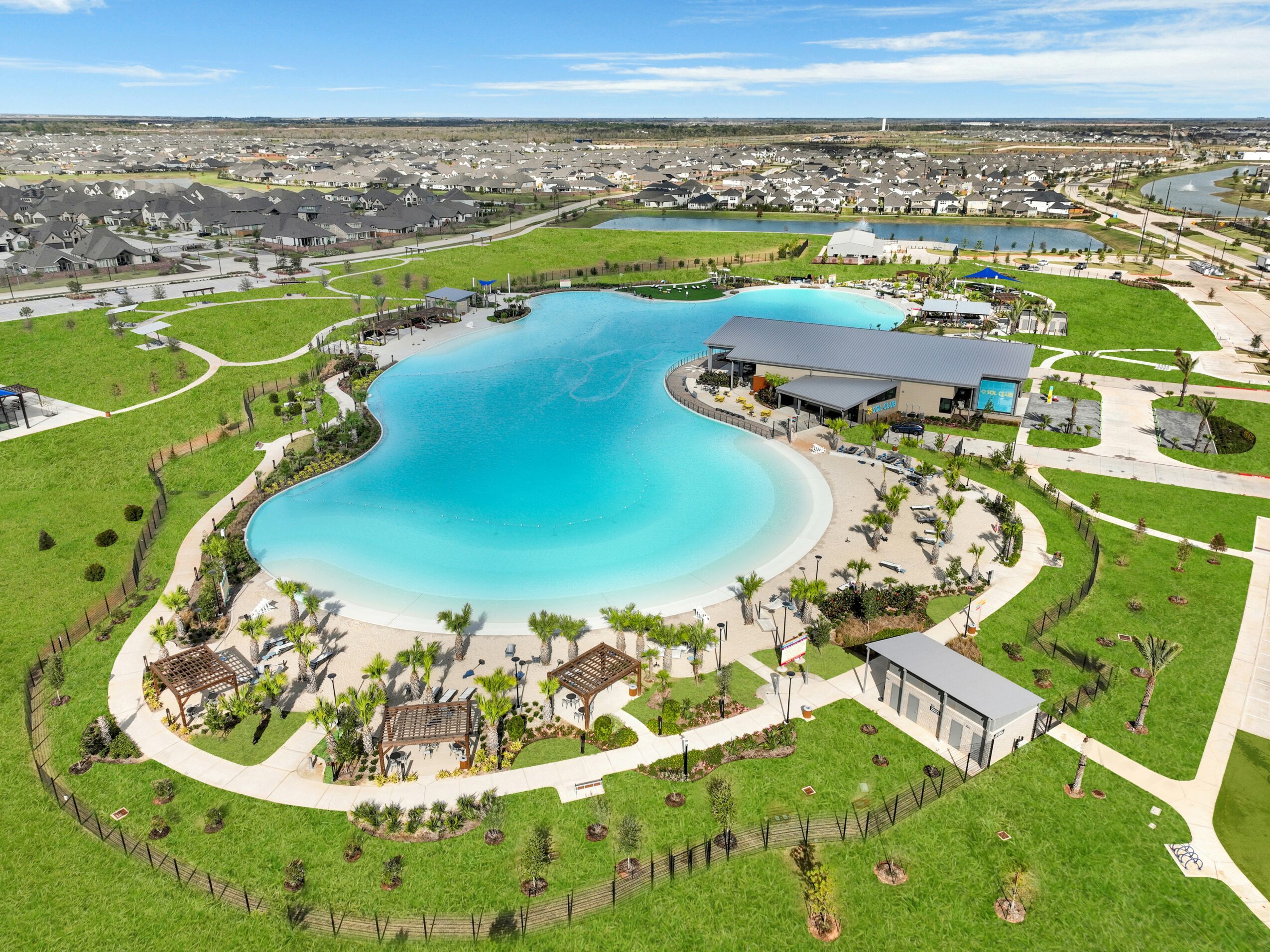 A large park with a central lake, walking paths, green lawns, and small structures, under a blue sky.