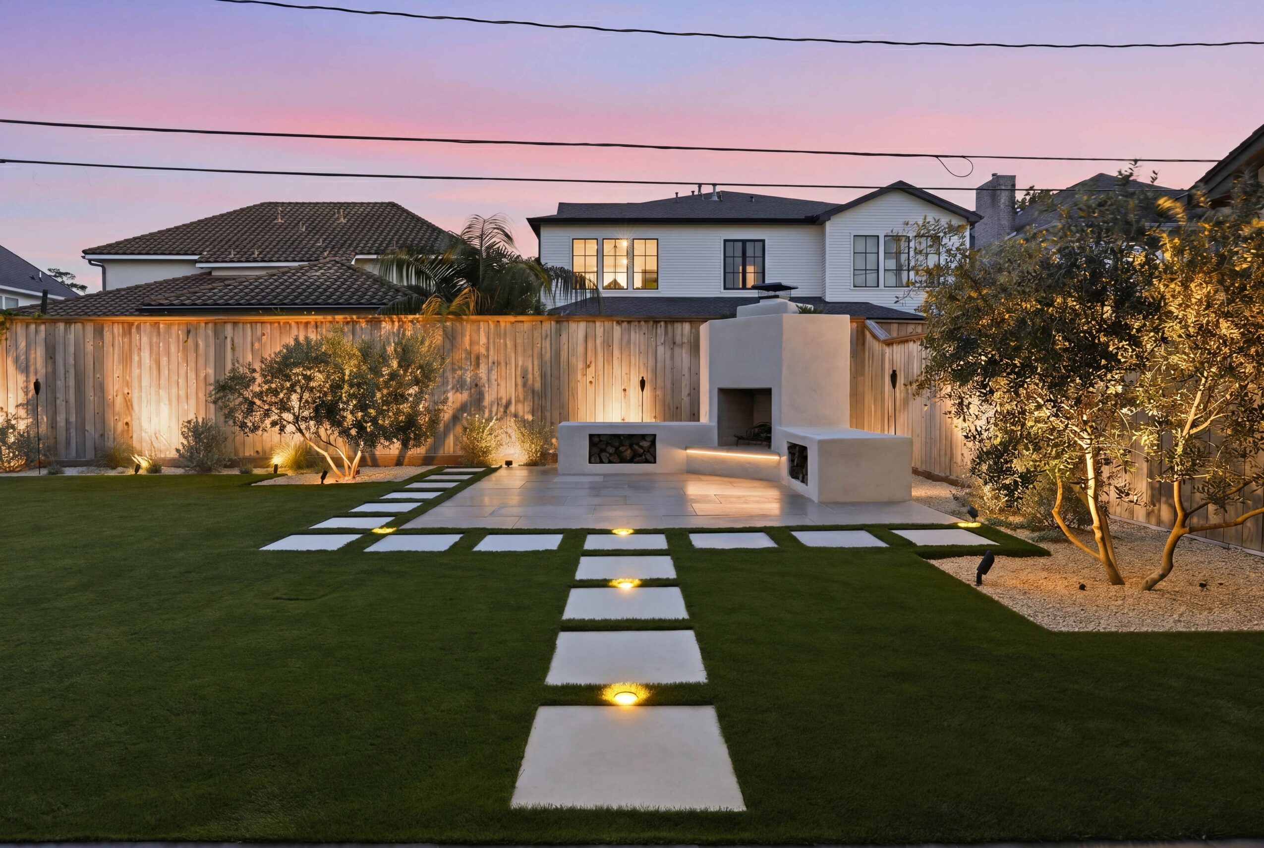 Modern backyard with pathway, grass, trees, and a house in the background during sunset.