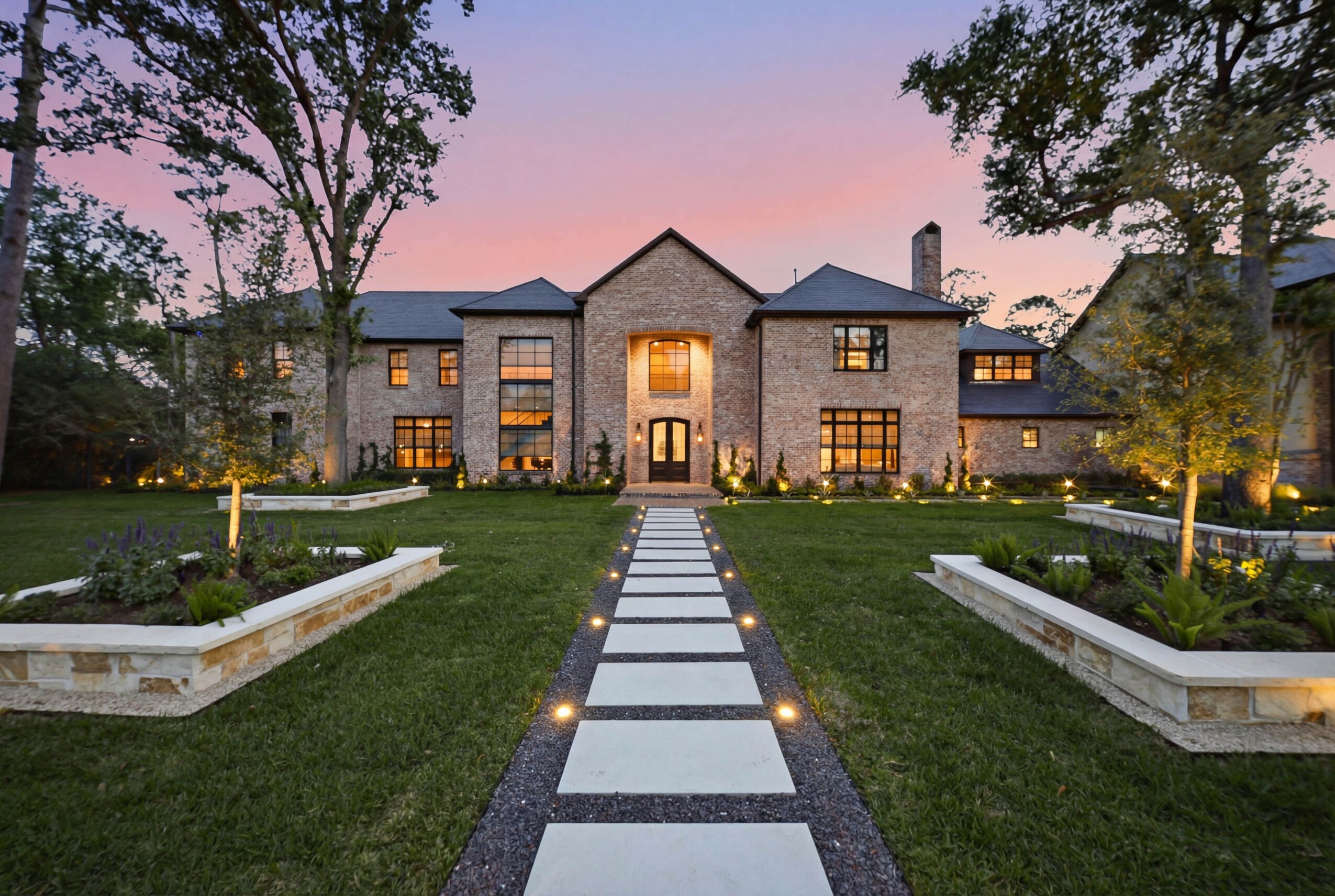 Large stone mansion with symmetrical windows and a central entrance, surrounded by a landscaped garden at sunset.