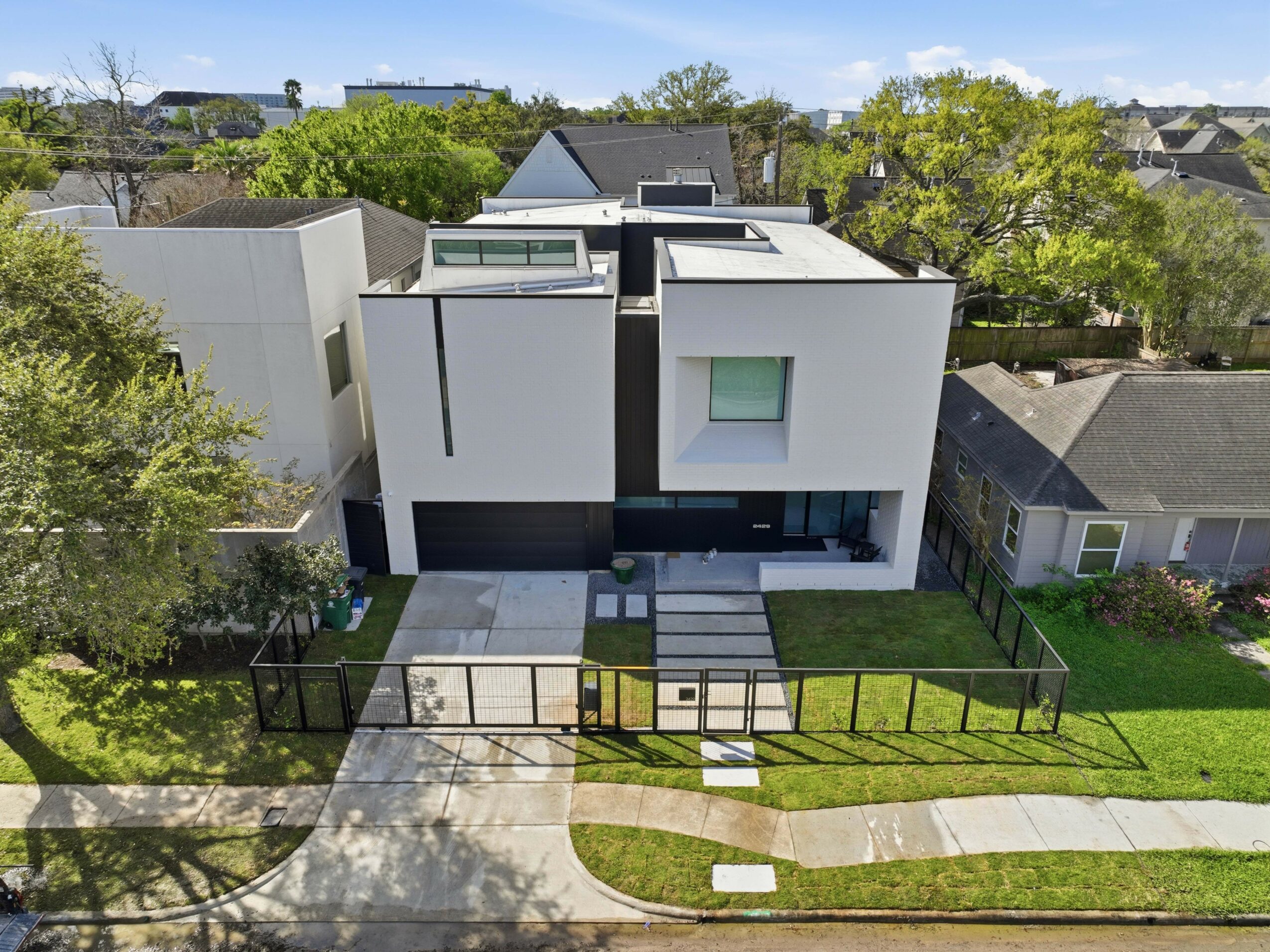 Modern white house with large windows, surrounded by green lawn and trees, fenced yard, and neighboring houses.
