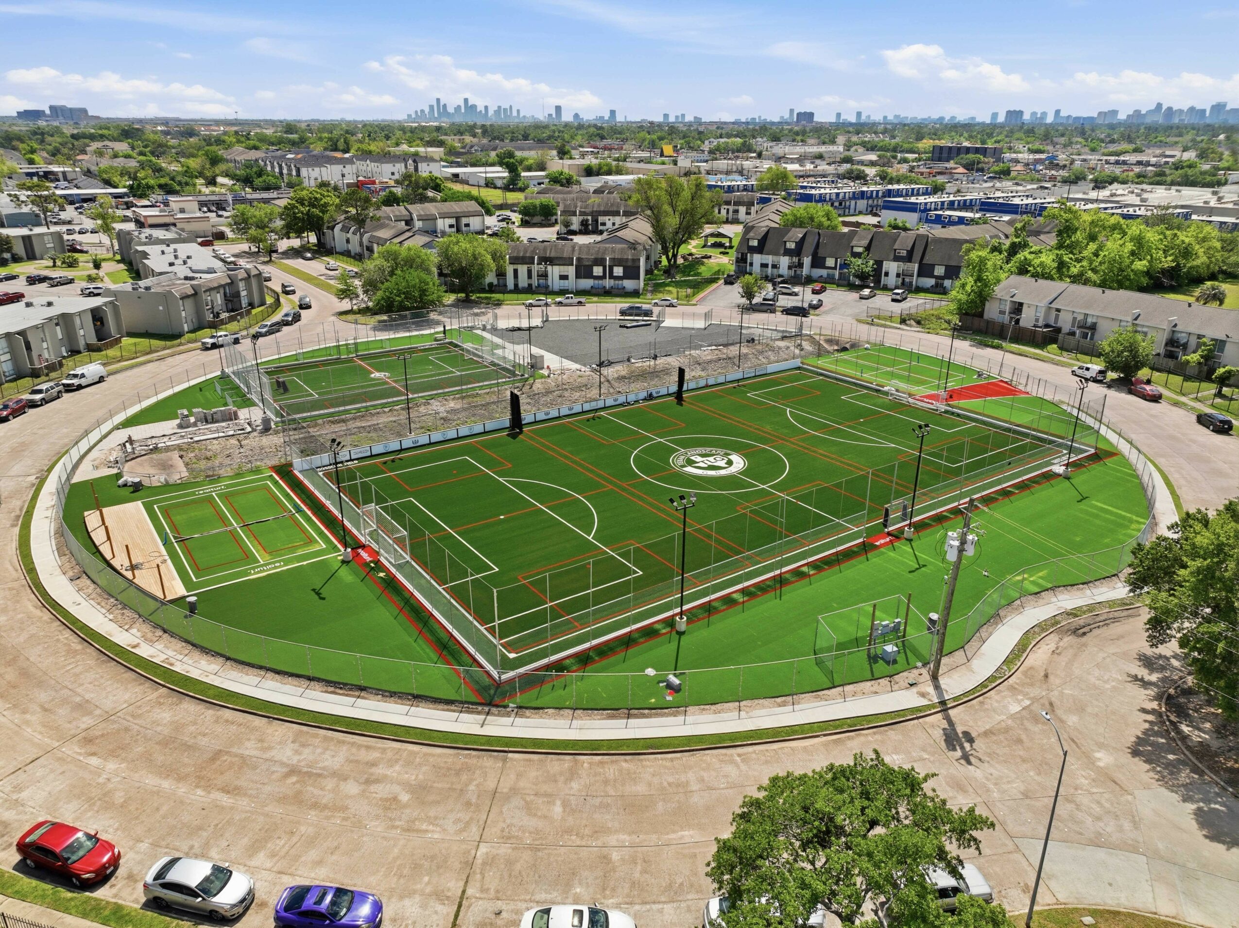 A sports field with multiple courts, surrounded by a running track and residential buildings in the background.