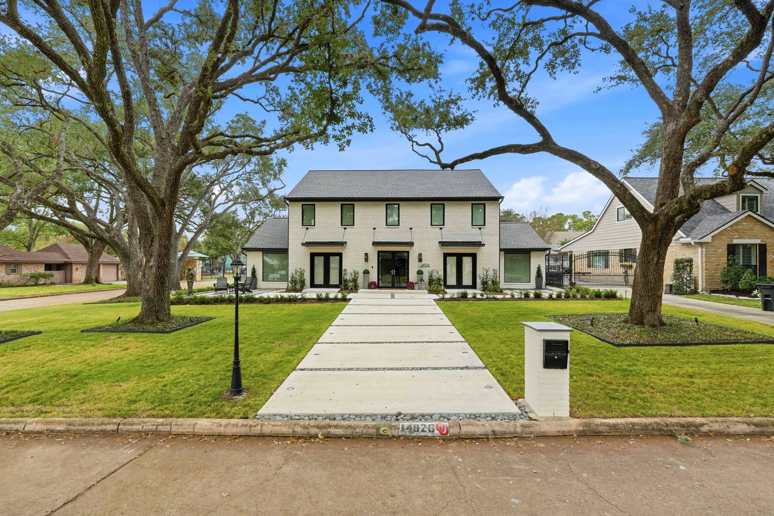 Two large trees frame a white house with a front porch, green lawn, and a paved walkway leading to the entrance.
