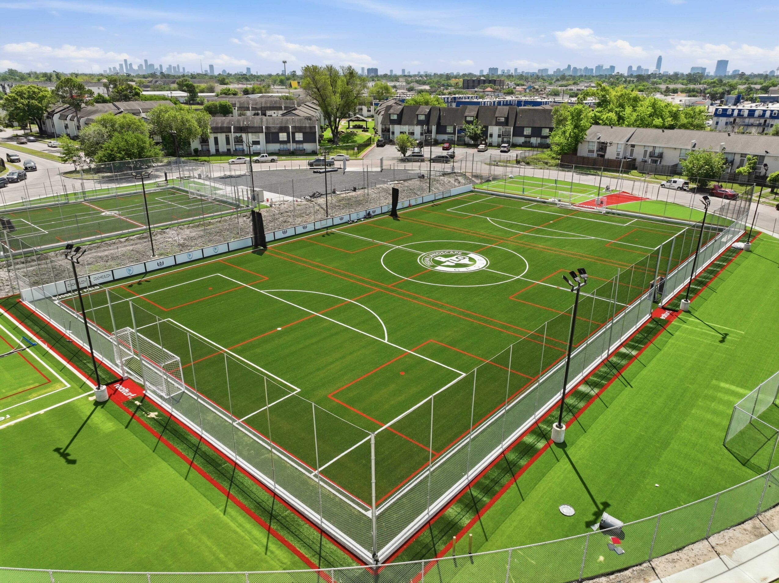Sports field with soccer and tennis courts, surrounded by a fence, under a blue sky with clouds.