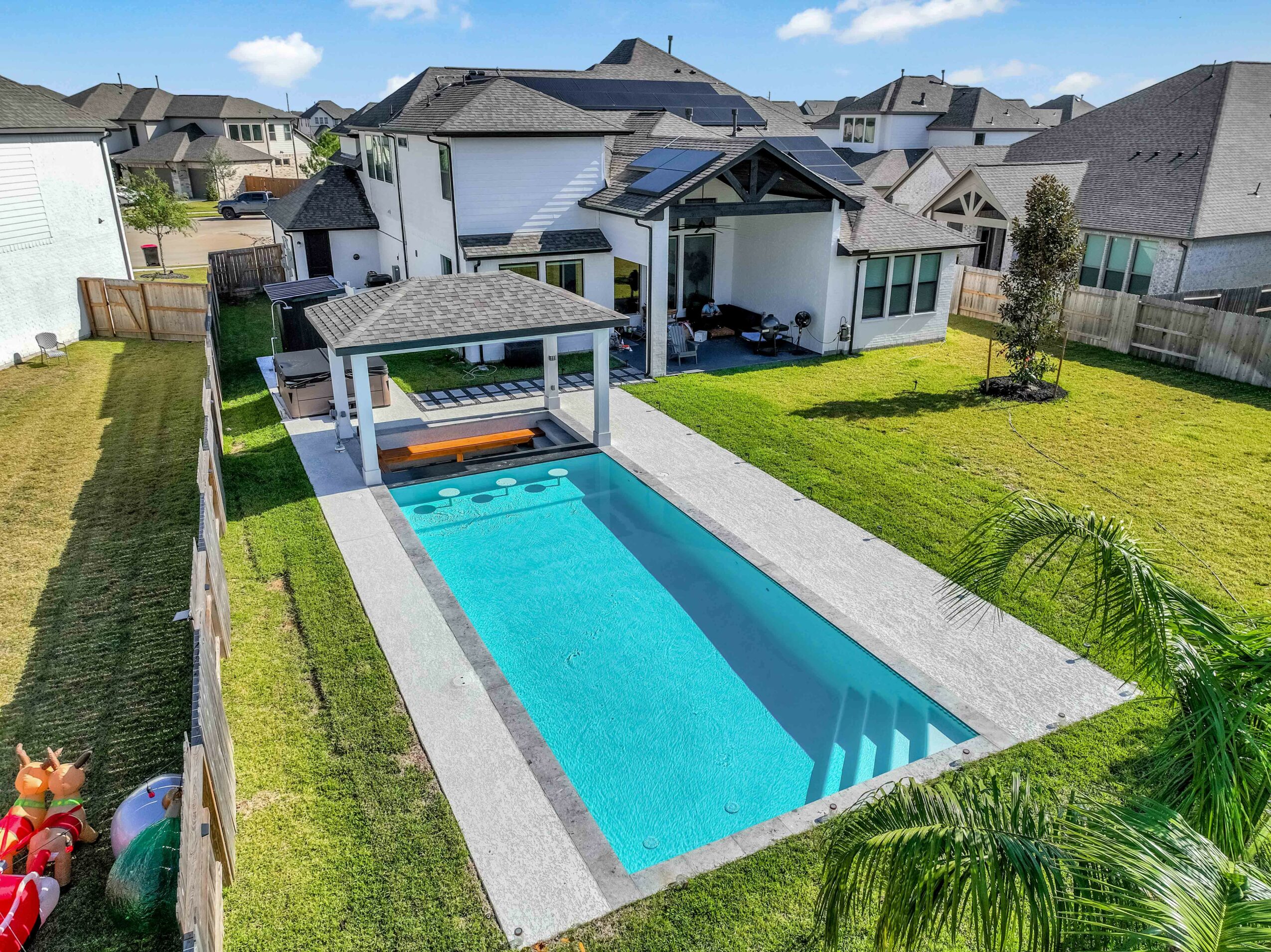 Backyard with a rectangular swimming pool, covered patio, green lawn, and neighboring houses in the background.