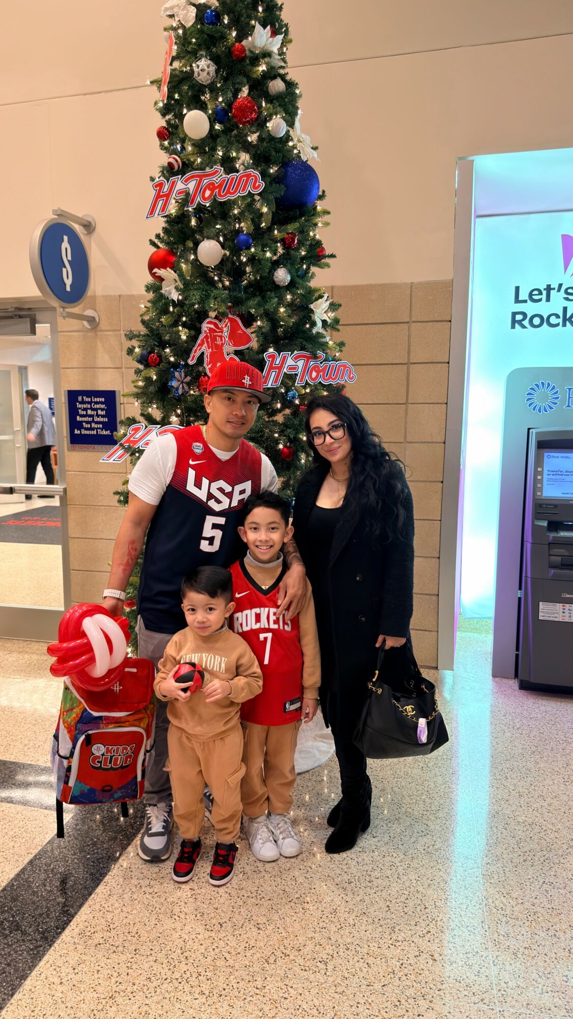 Family of three children and an adult woman standing in front of a decorated Christmas tree at an indoor location.