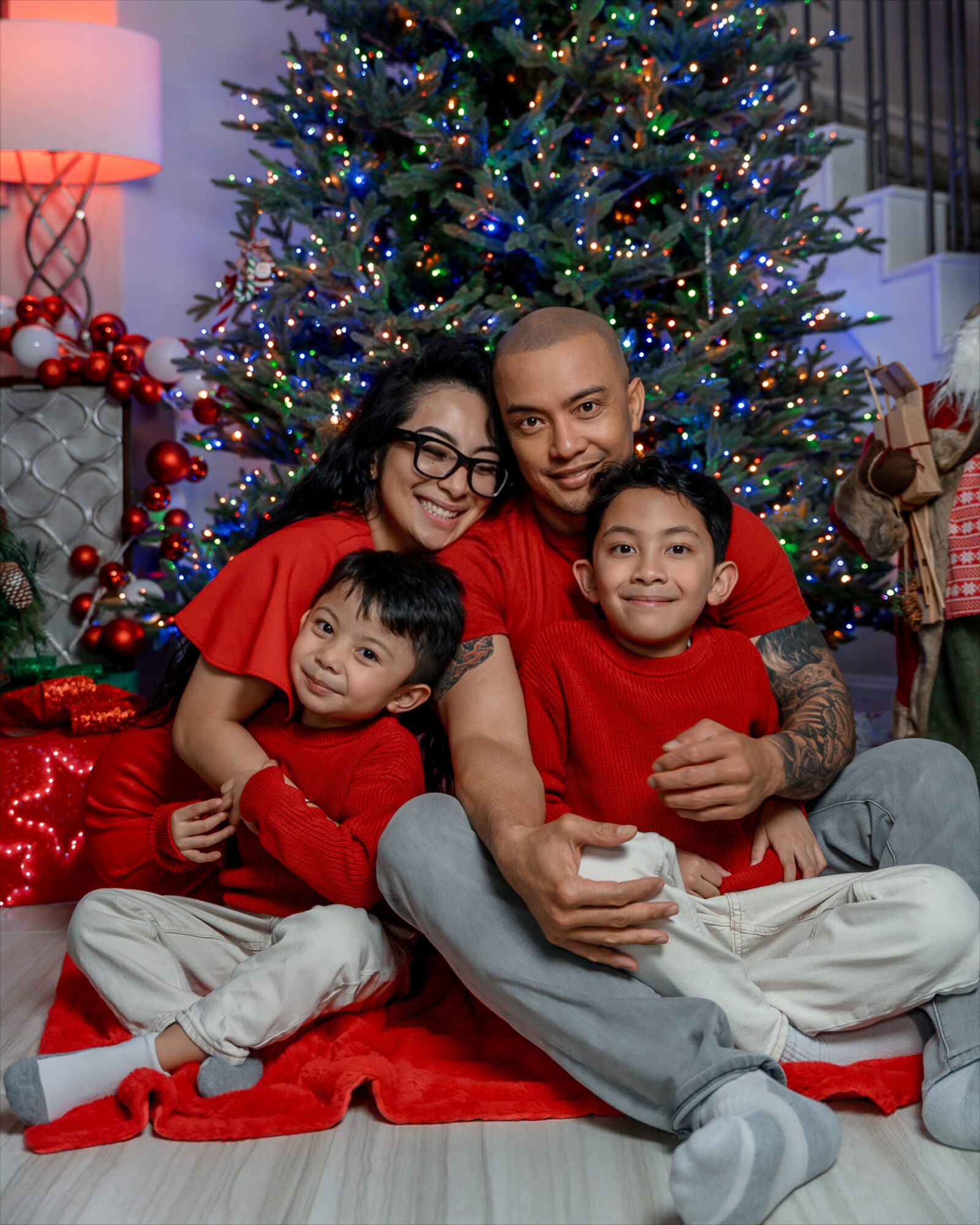 Family of four sitting on the floor in front of a decorated Christmas tree, smiling, wearing red clothes.