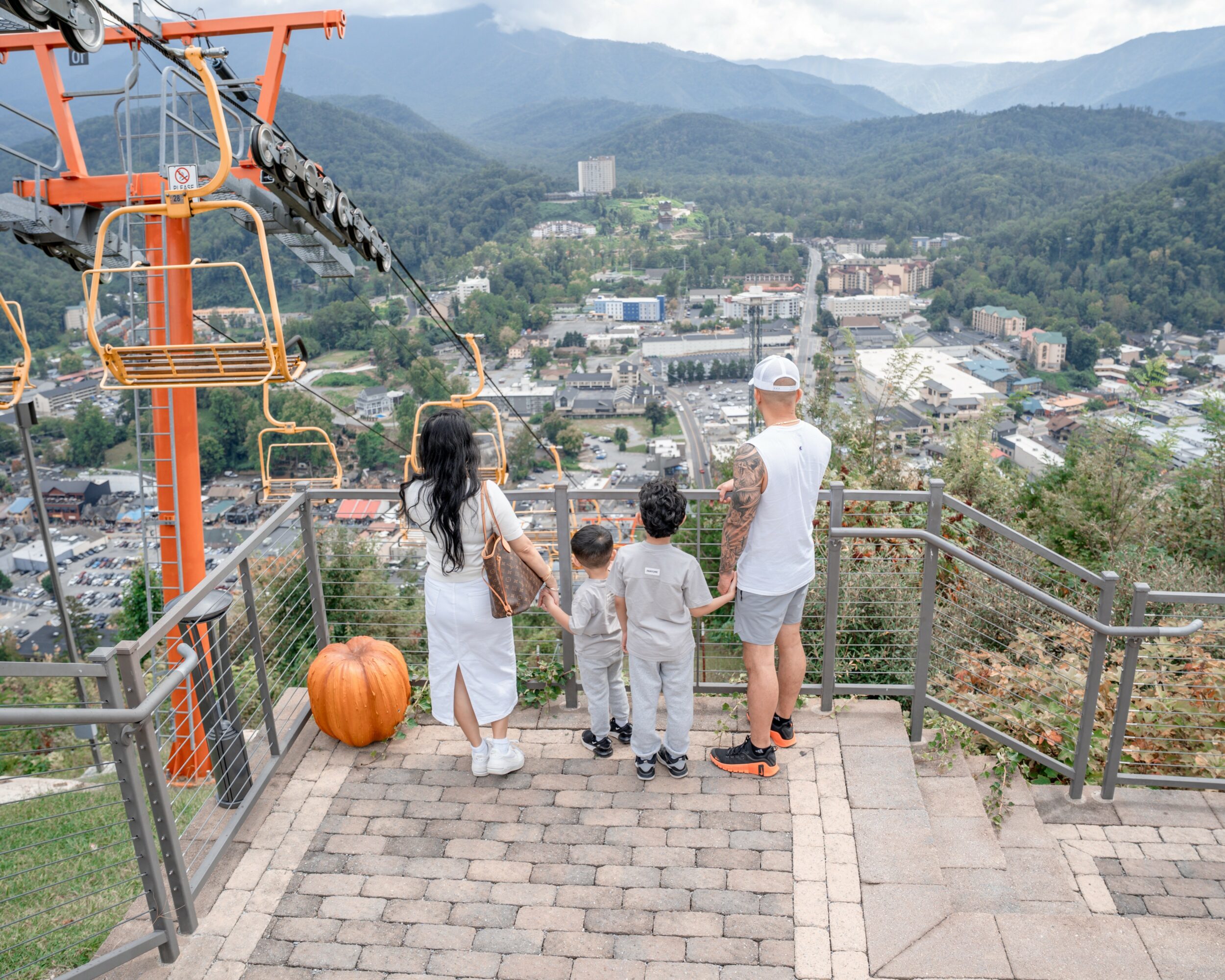 Four people, two children and two adults, stand on a viewing platform overlooking a town and mountains, with a pumpkin nearby.