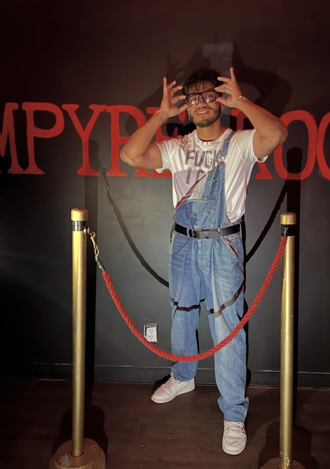 Person standing behind a red rope barrier, making hand gestures, in front of a dark background with red text.