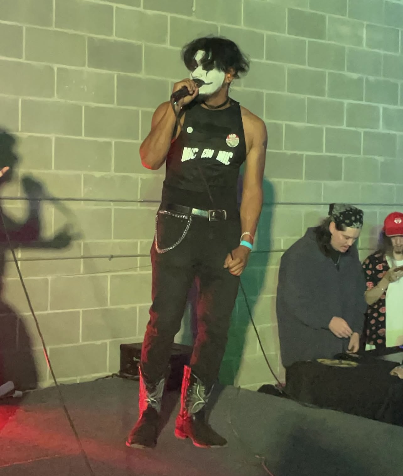 Person with black curly hair, wearing a sleeveless black shirt, holding a microphone, standing against a brick wall.