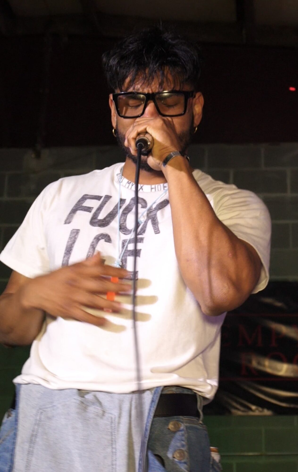 Person singing into microphone, wearing glasses and a white T-shirt, in an indoor setting.