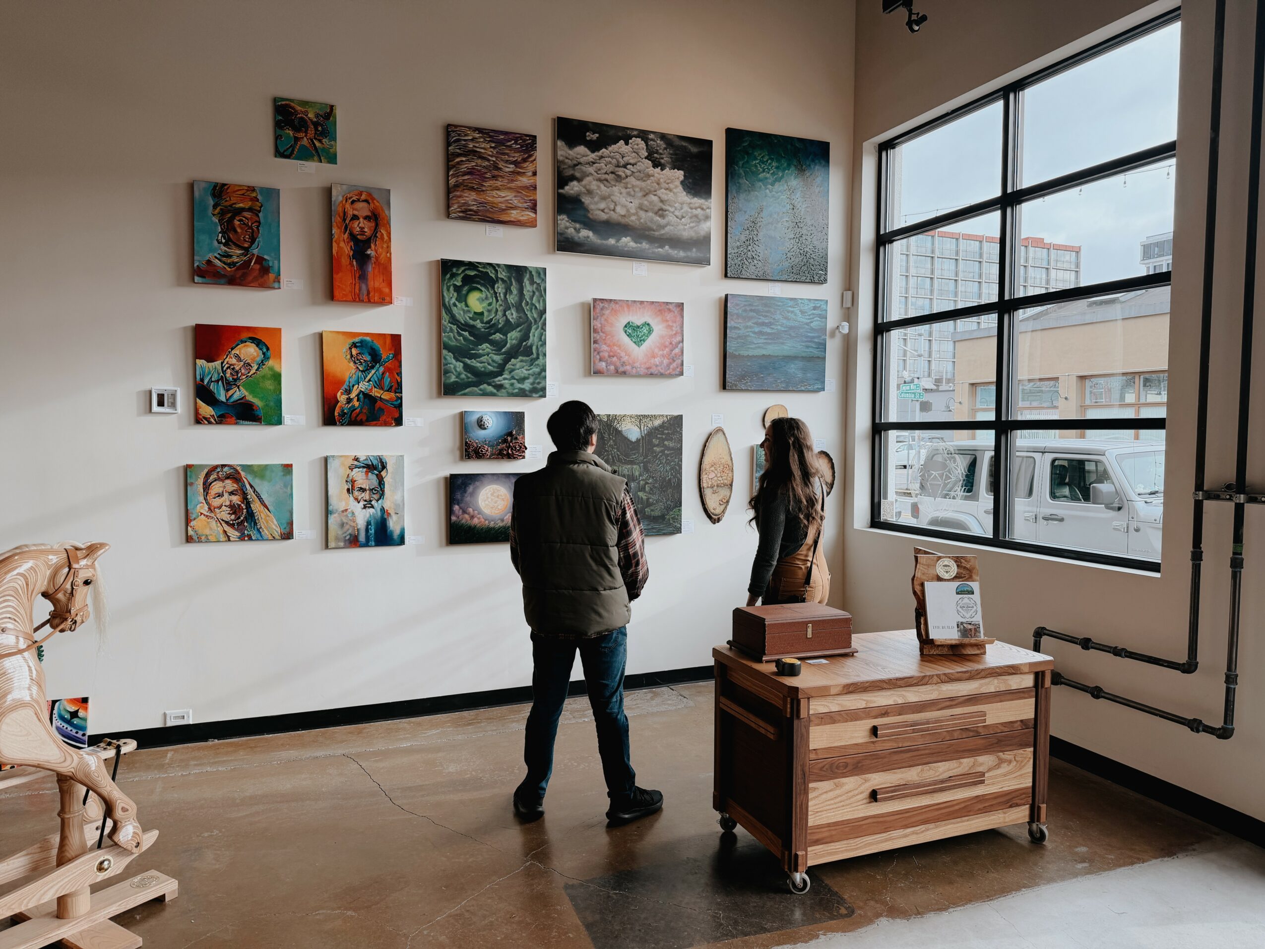 Two people stand in an art gallery looking at various paintings on the wall, with large windows showing city buildings outside.