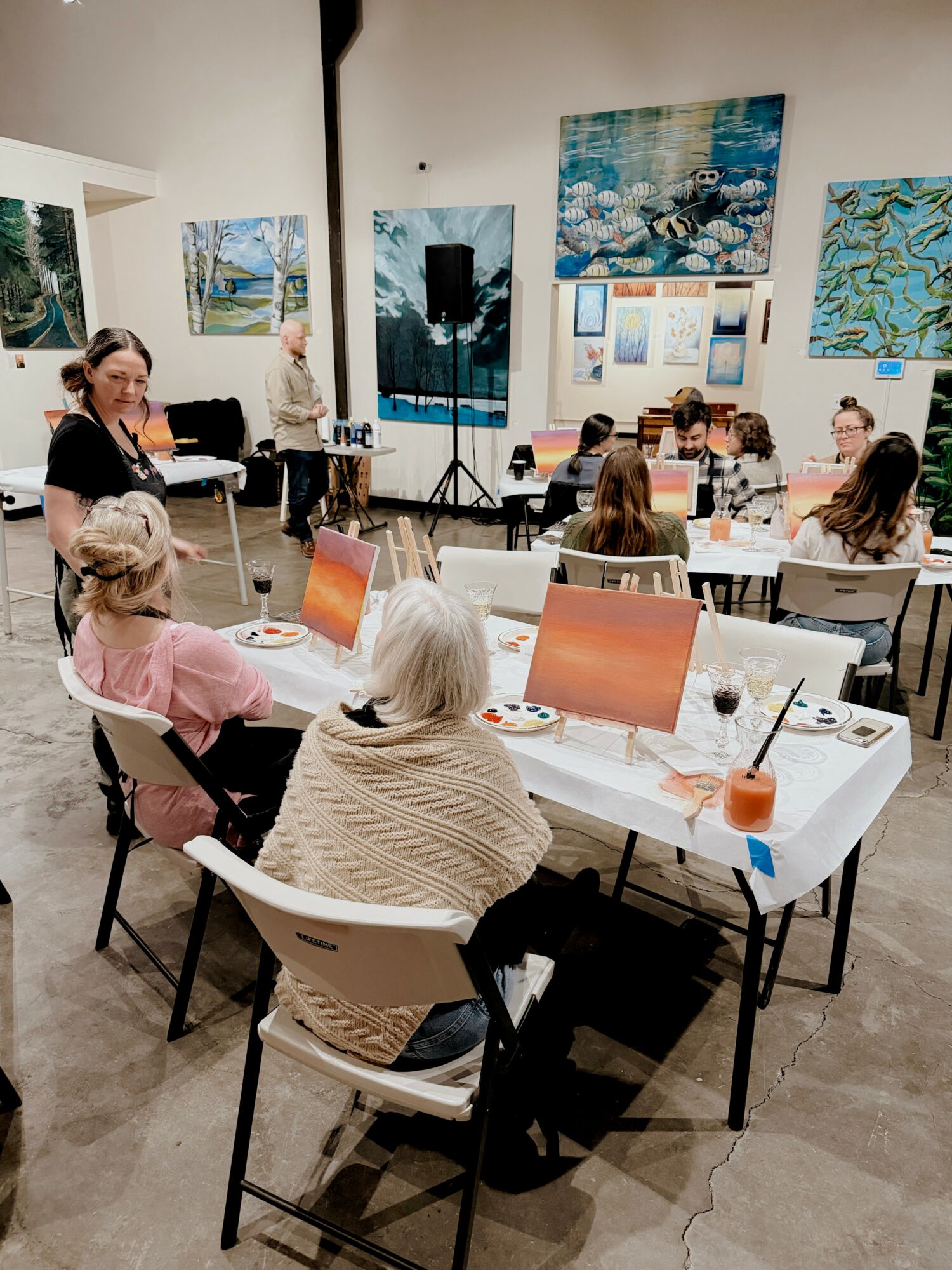 People seated at tables in an art gallery, with paintings on the walls, some people taking notes or listening.