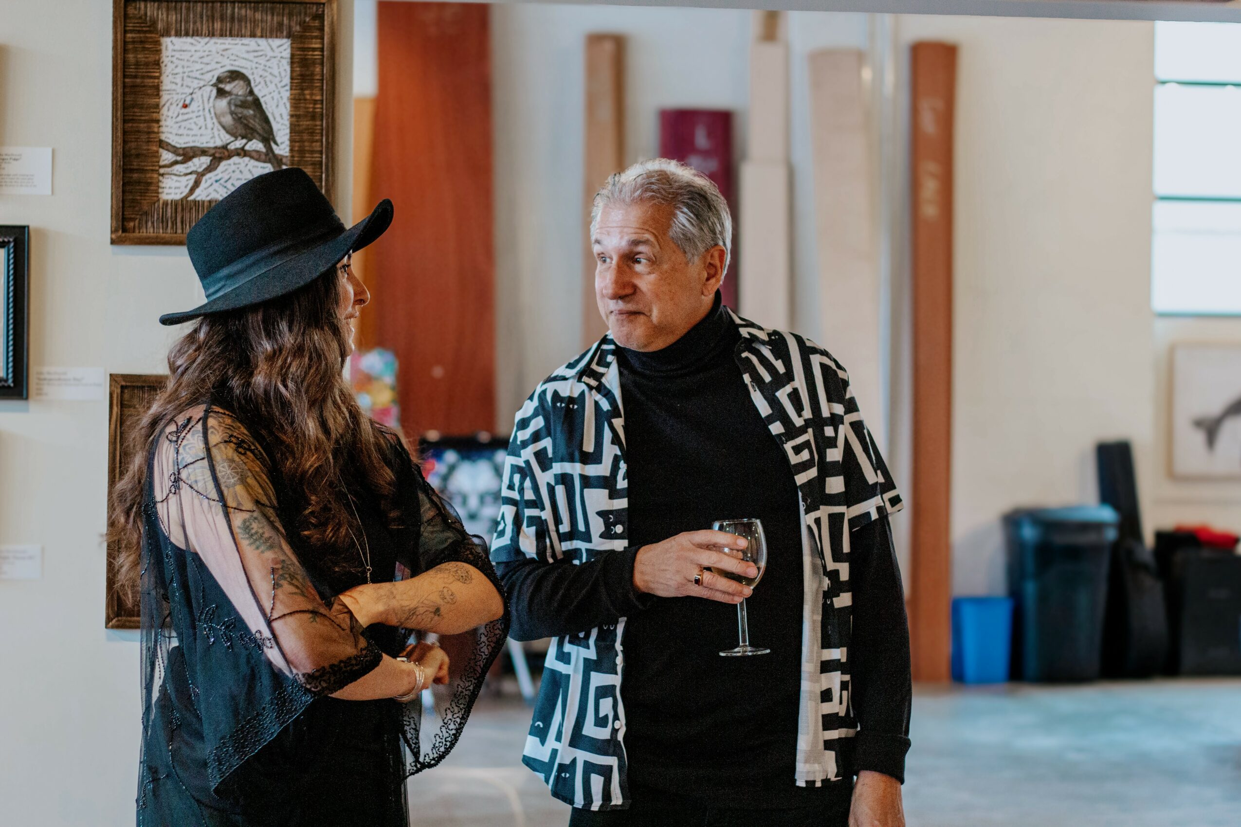 Two people talking indoors, one holding a glass of wine, with artwork and furniture in the background.