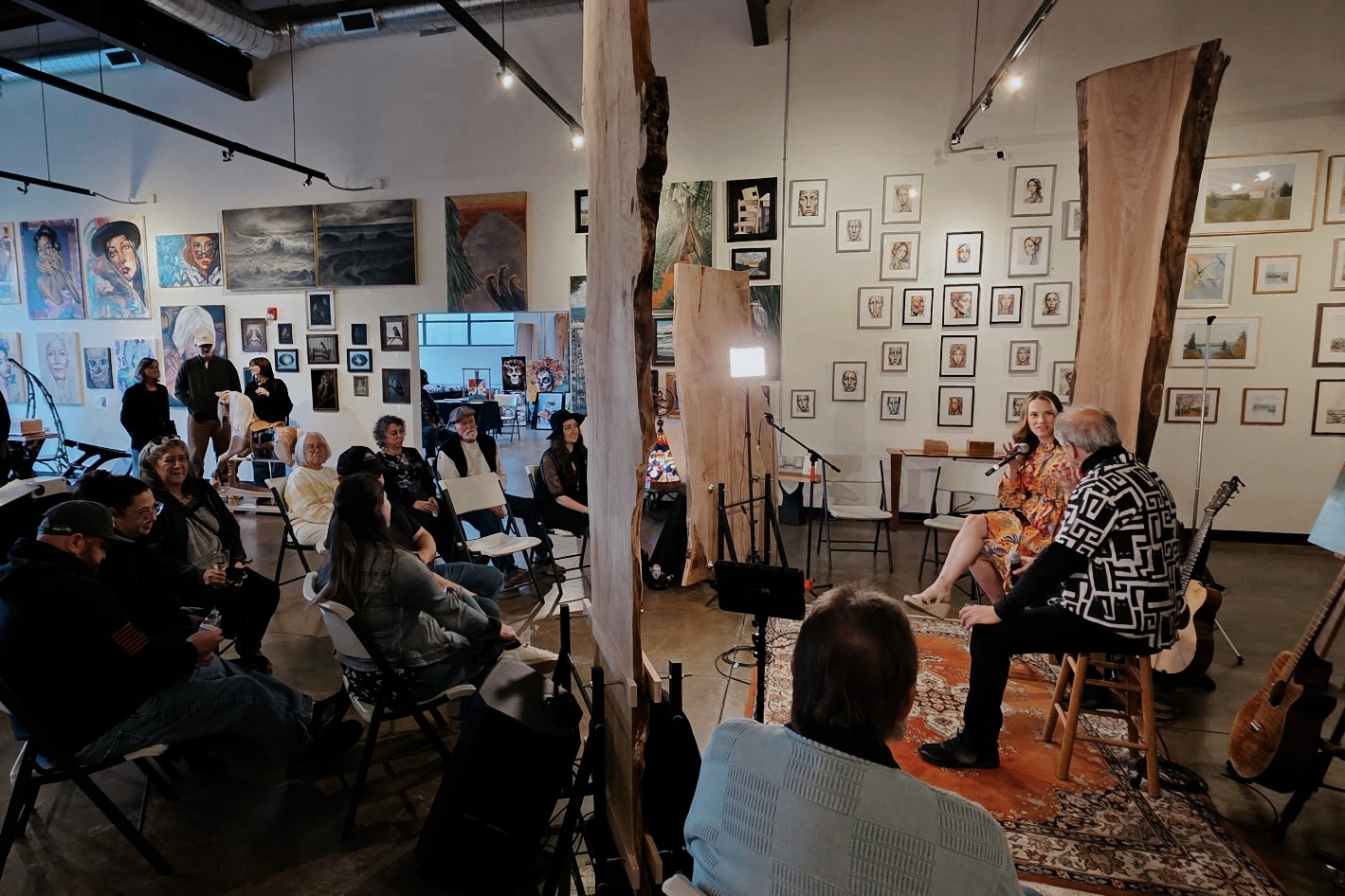 People seated in an art gallery listening to two women speaking on a stage, surrounded by framed artwork on walls.