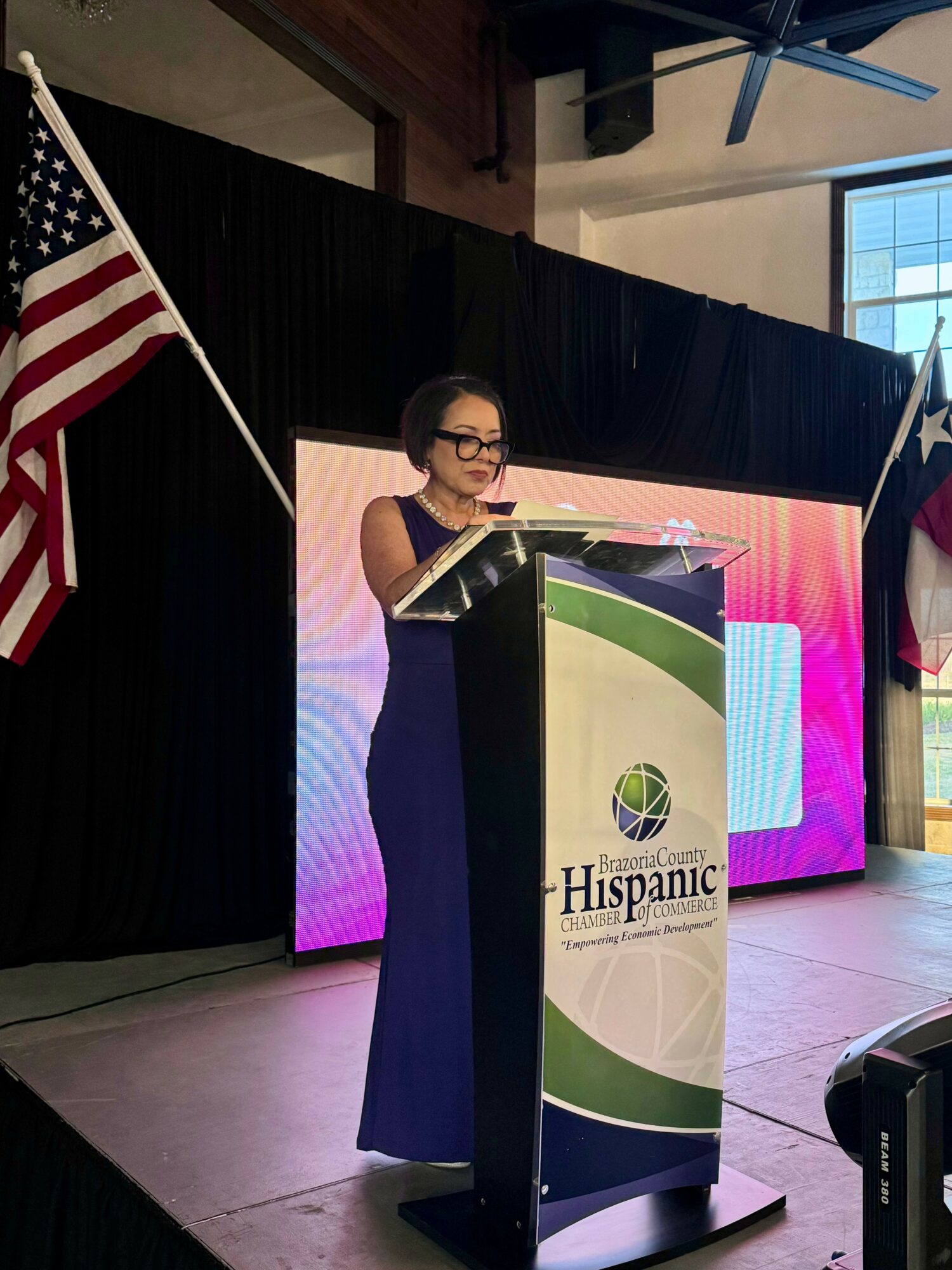 Woman in a dark dress and glasses speaking at a podium with a Hispanic-themed logo, American flags on stage.