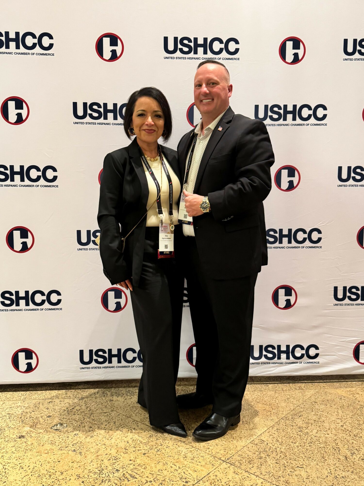 Two people standing together in front of a backdrop with USHCC logos, smiling, holding drinks, dressed professionally.