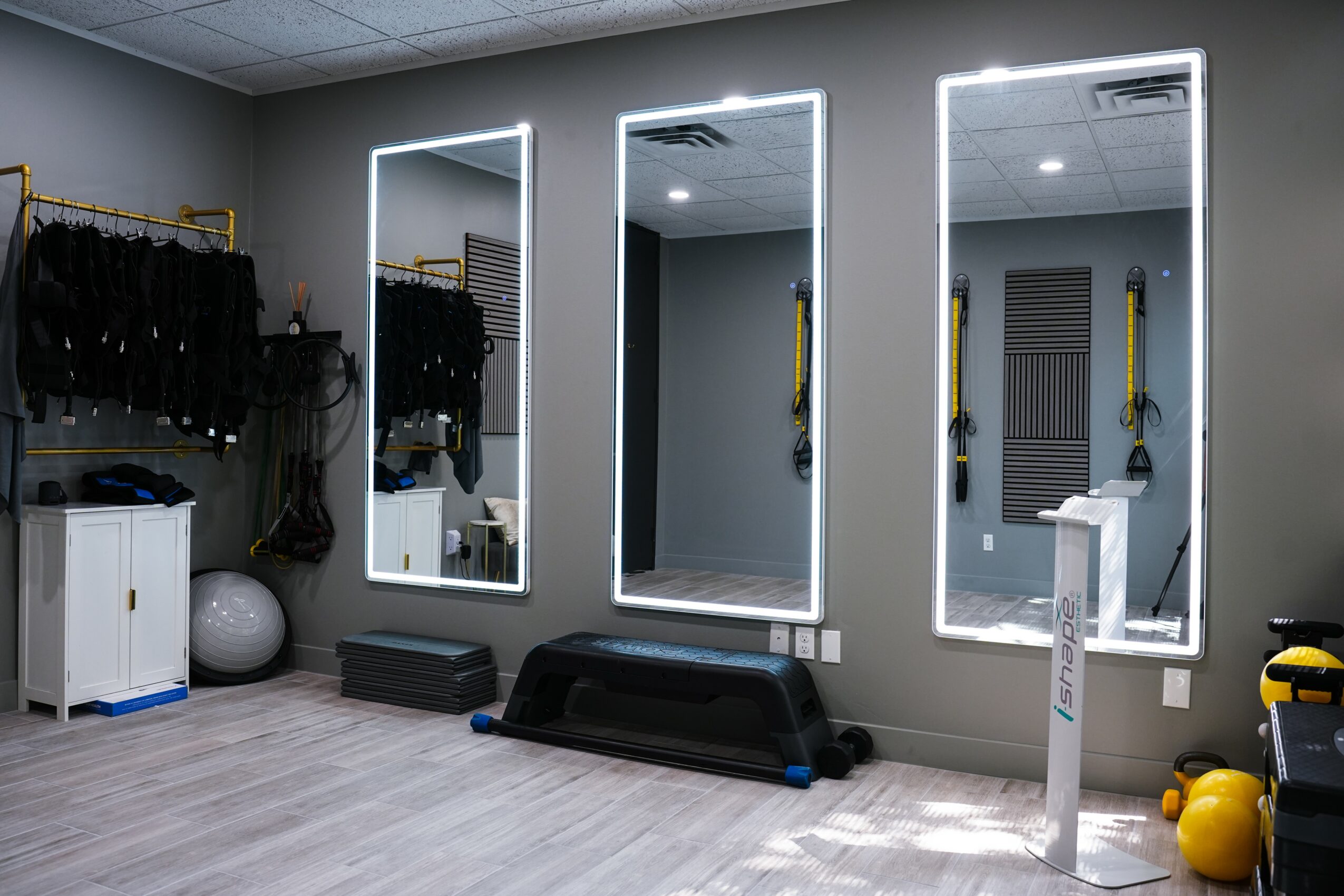 Gym with three illuminated mirrors on a gray wall, exercise equipment, and a white cabinet with black clothing. Light wood flooring.