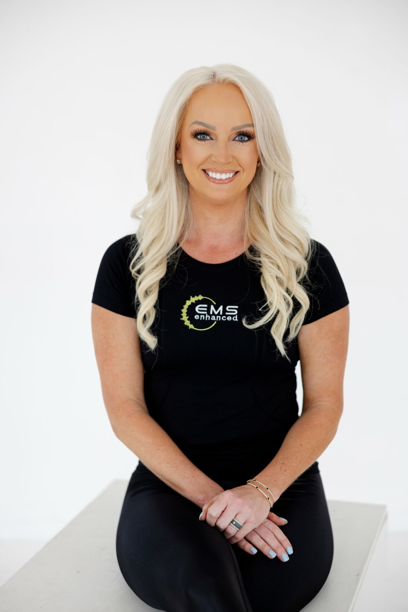 Smiling woman with long blonde hair, wearing a black t-shirt with a logo, sitting on a white surface against a plain background.