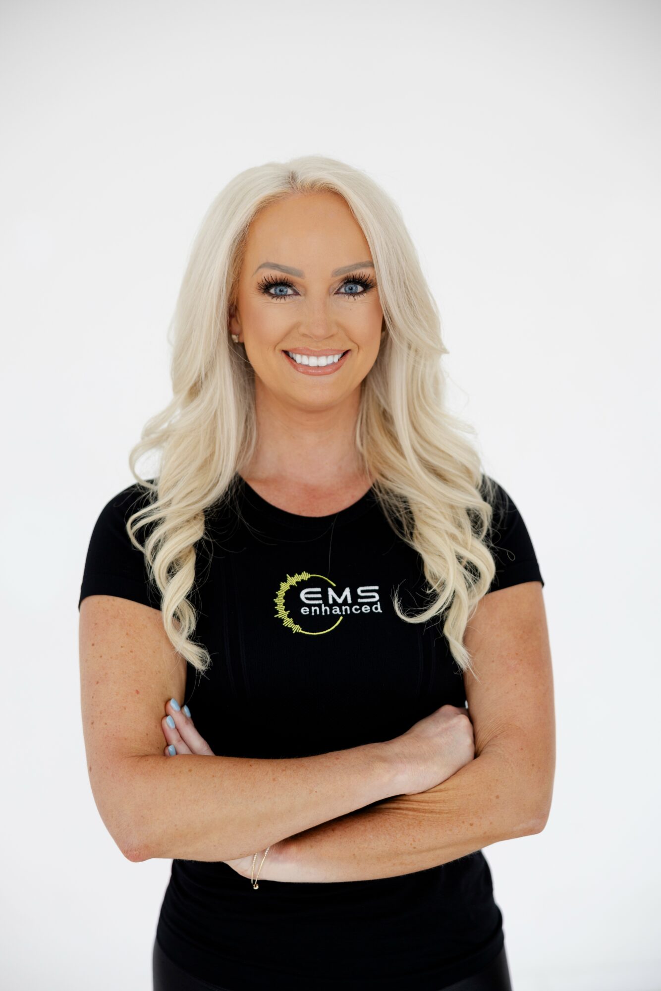 Smiling woman with long blonde hair wearing a black EMS Academy t-shirt, arms crossed, white background.