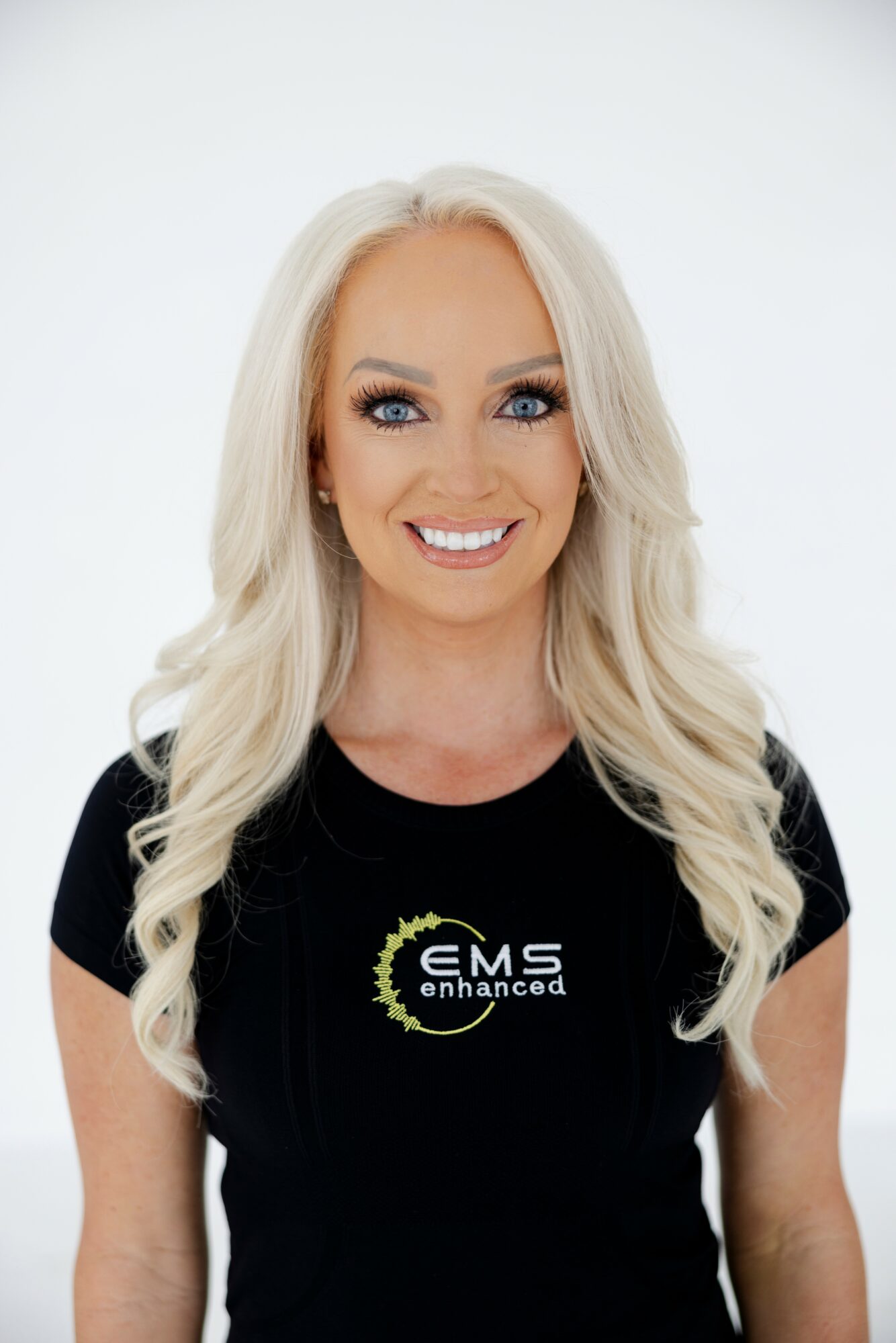 Woman with long blonde hair smiling, wearing a black shirt with EMS logo, against white background.