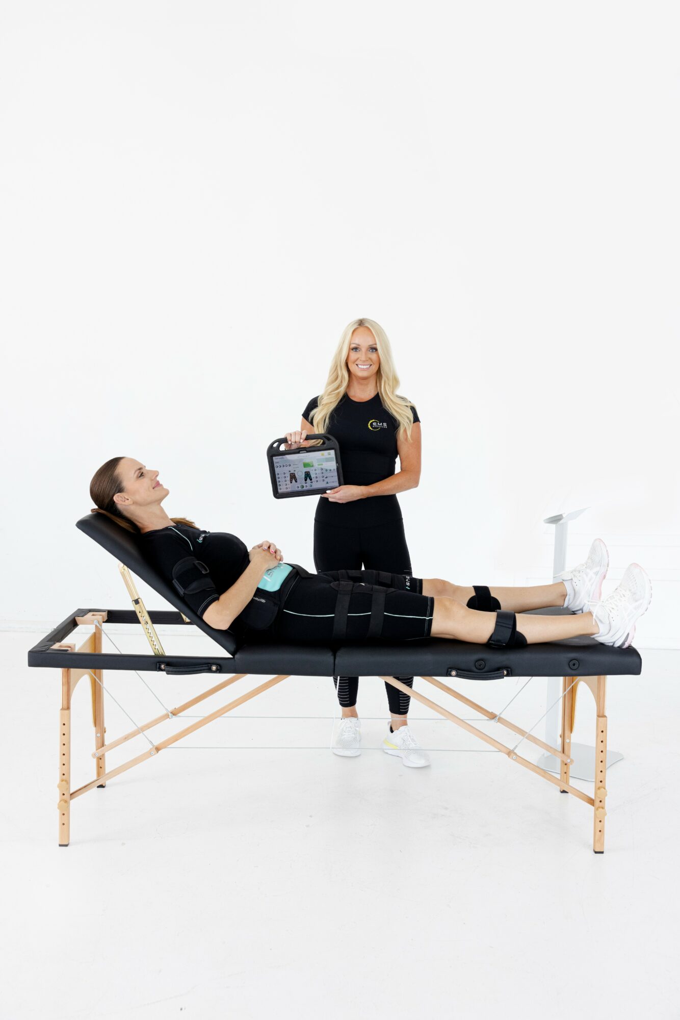 Woman lying on examination table with bandage on abdomen, another woman standing holding a tablet, white background.