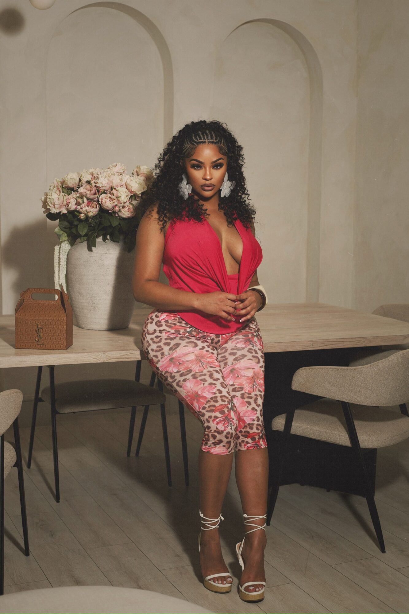 Woman in red top and pink patterned skirt standing by a table with flowers and a chair.