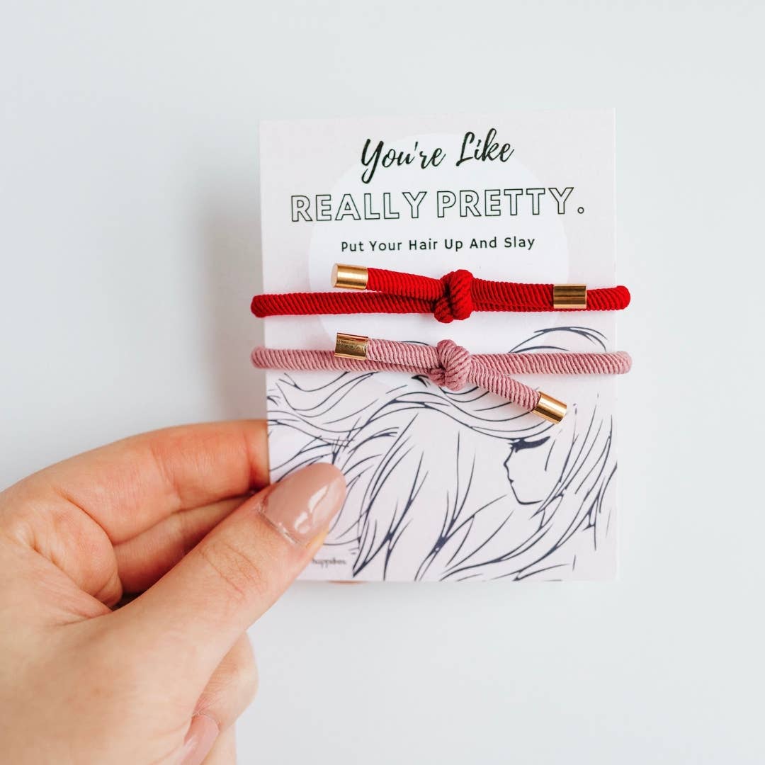 Hand holding a card with two pink and red braided bracelets, background is white with black line drawing of a person's face.