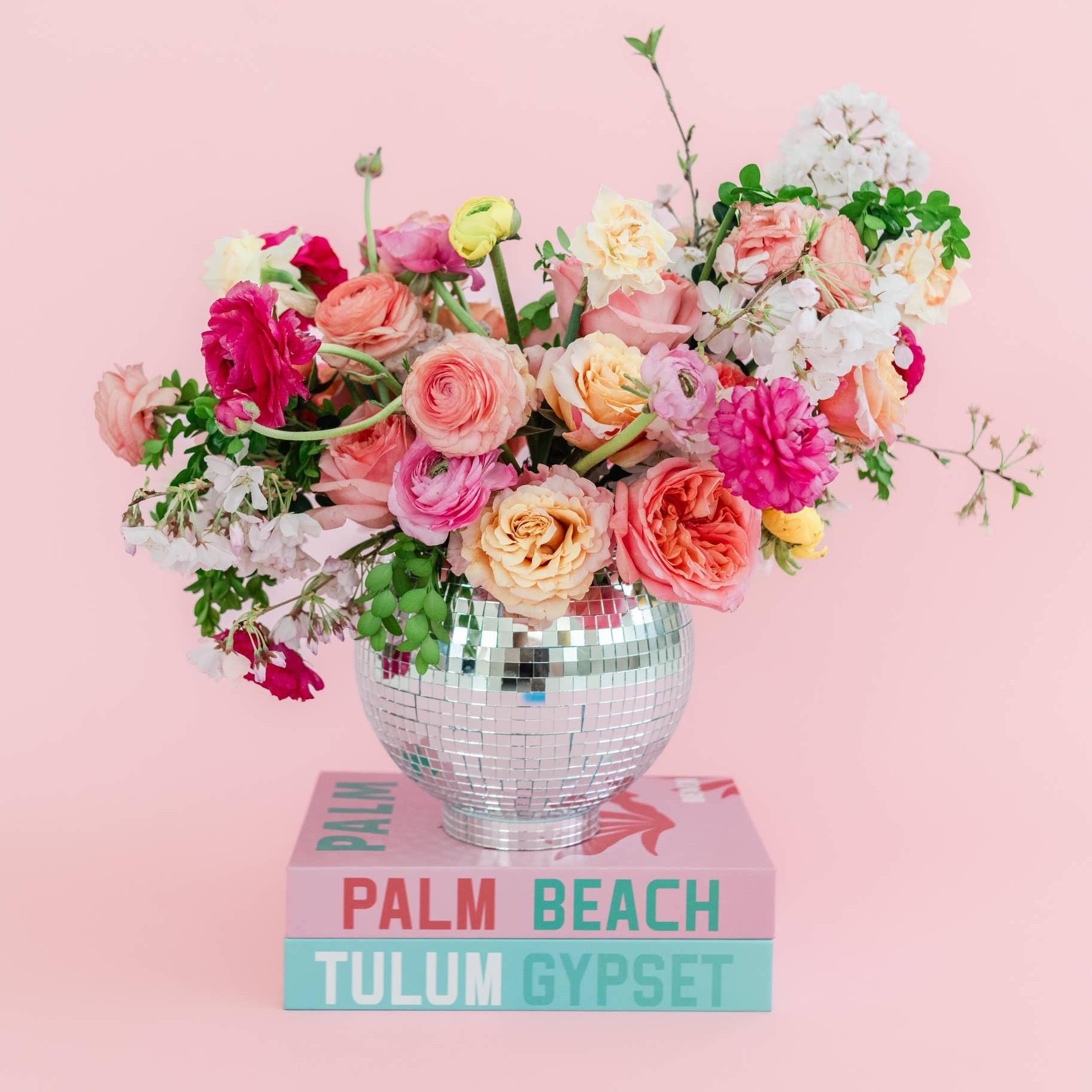 Colorful bouquet of pink, white, and yellow flowers in a reflective silver vase on stacked books against pink background.