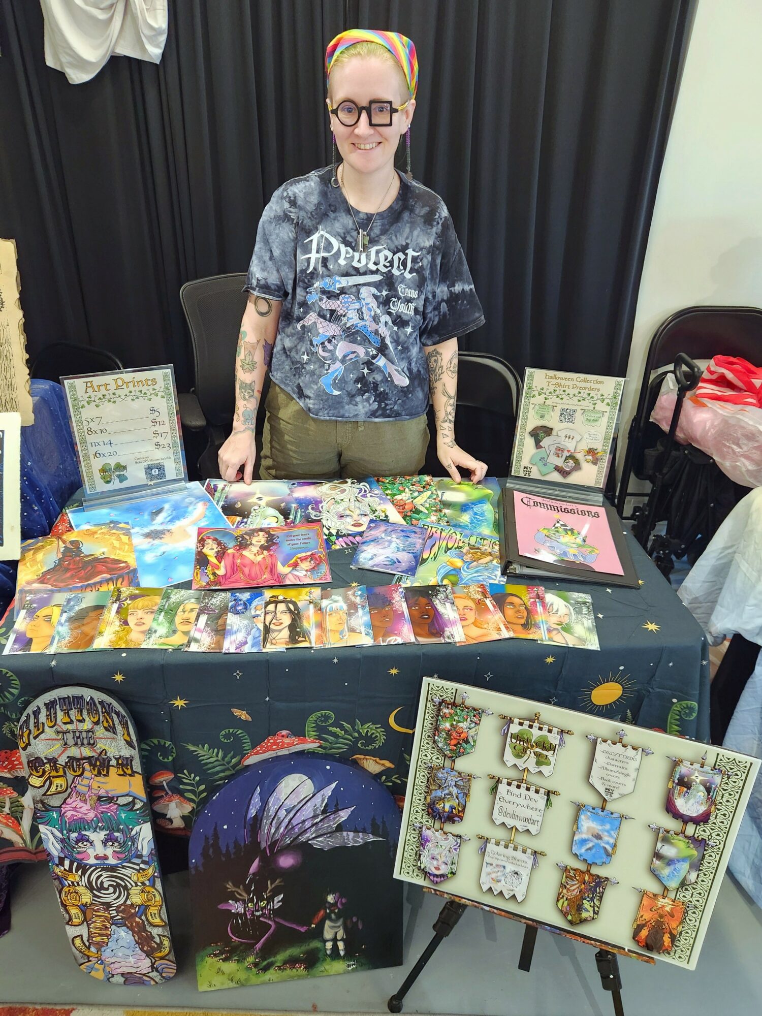 Person with glasses and colorful headscarf standing behind a table with colorful artwork and crafts, black curtain background.